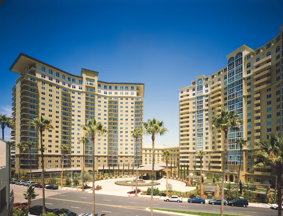 Exterior view of a large, modern senior living facility with two tall, curved buildings surrounding a circular driveway lined with palm trees under a clear blue sky.