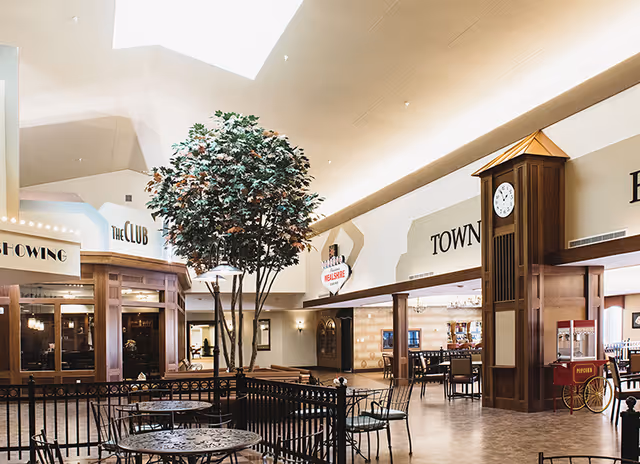 Interior view of a senior living facility featuring a spacious common area with a tall artificial tree in the center, metal tables and chairs arranged around it, a large wooden clock tower, and various signs on the walls indicating areas such as The Club, Town Hall, and Wellness Center.