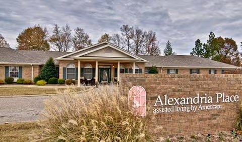 Exterior view of Alexandria Place assisted living facility showing a single-story brick building with a covered entrance, surrounded by trees and landscaping under a cloudy sky. A brick sign in the foreground displays the facility name and logo.
