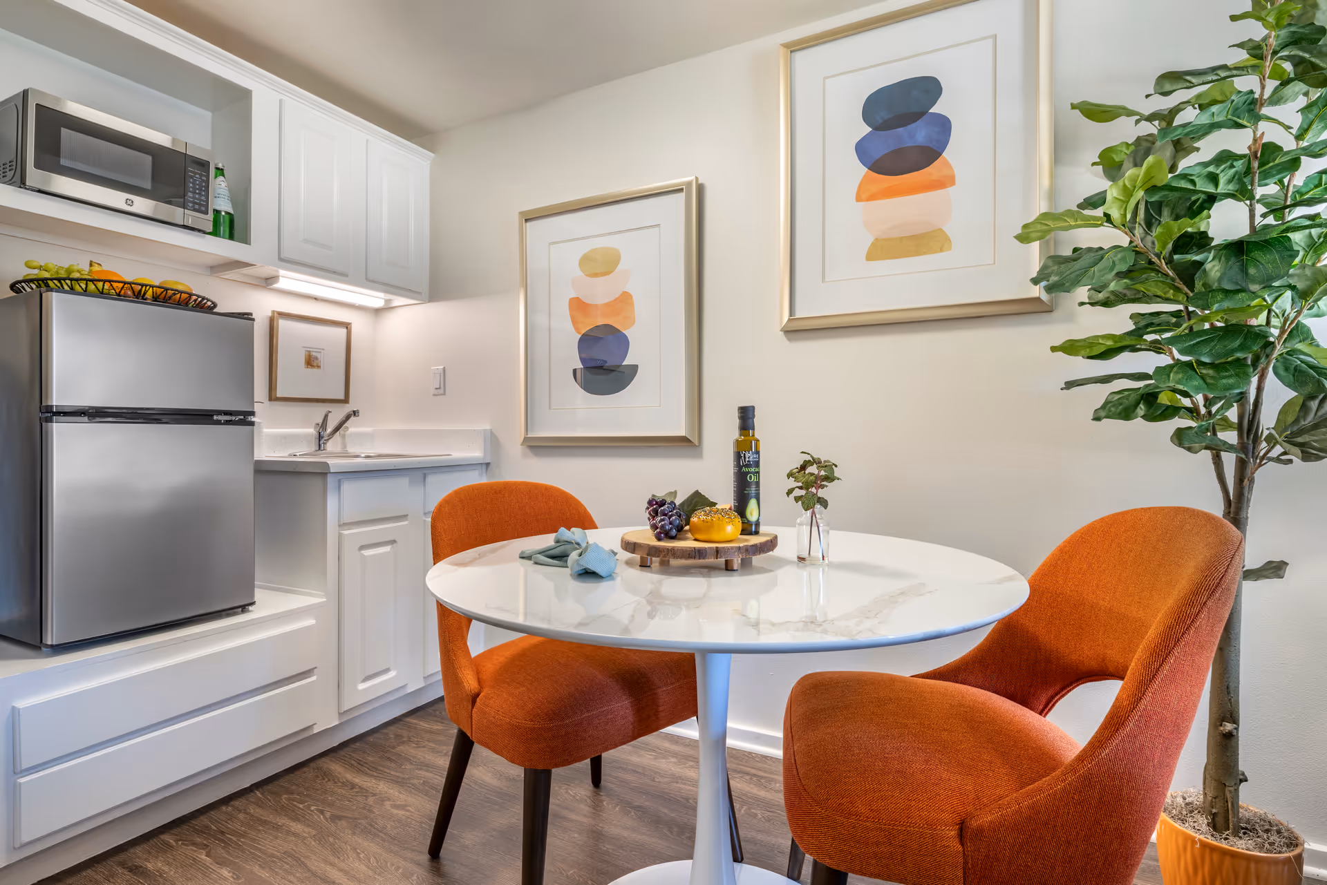 A small dining area with a round white marble table and two orange upholstered chairs. On the table are a bottle of avocado oil, a small vase with a plant, grapes, and a yellow fruit on a wooden tray. Behind the table is a kitchenette with a stainless steel microwave and refrigerator, white cabinets, and a small sink. Two framed abstract art pieces hang on the wall, and a tall green potted plant is positioned to the right.