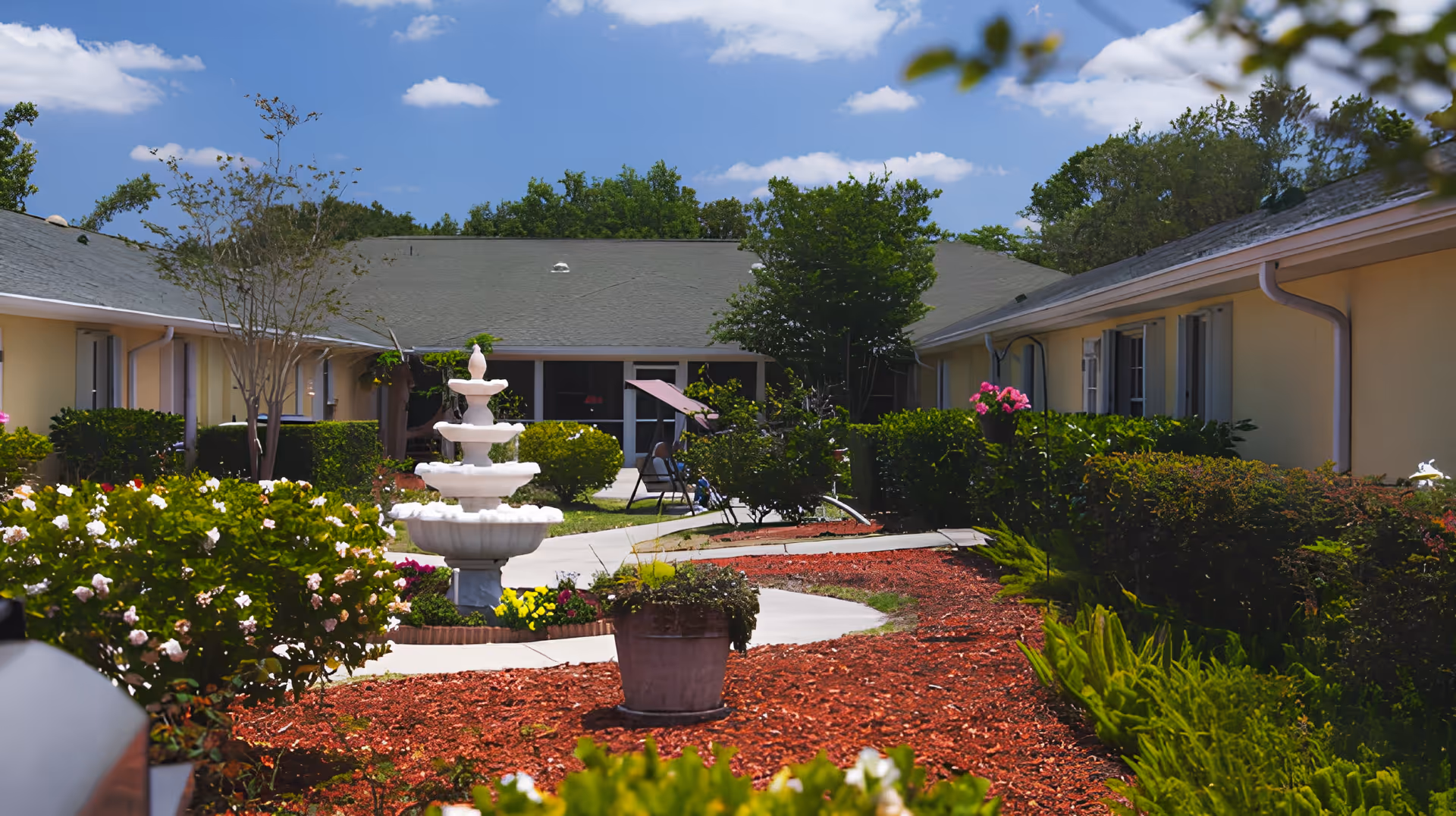 Outdoor courtyard area of a senior living facility with a white multi-tiered fountain, landscaped garden beds with flowers and shrubs, a curved concrete walkway, and single-story buildings with light yellow walls and gray roofs under a blue sky with scattered clouds.
