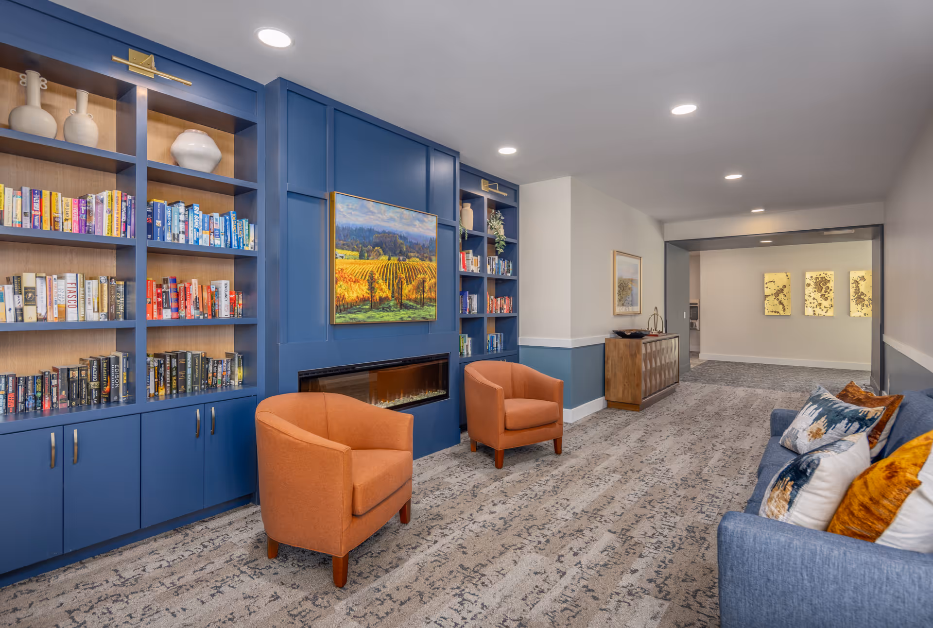 A bright lounge with blue built-in bookshelves, a wall-mounted TV above a fireplace, two orange armchairs, and a sofa with decorative pillows.