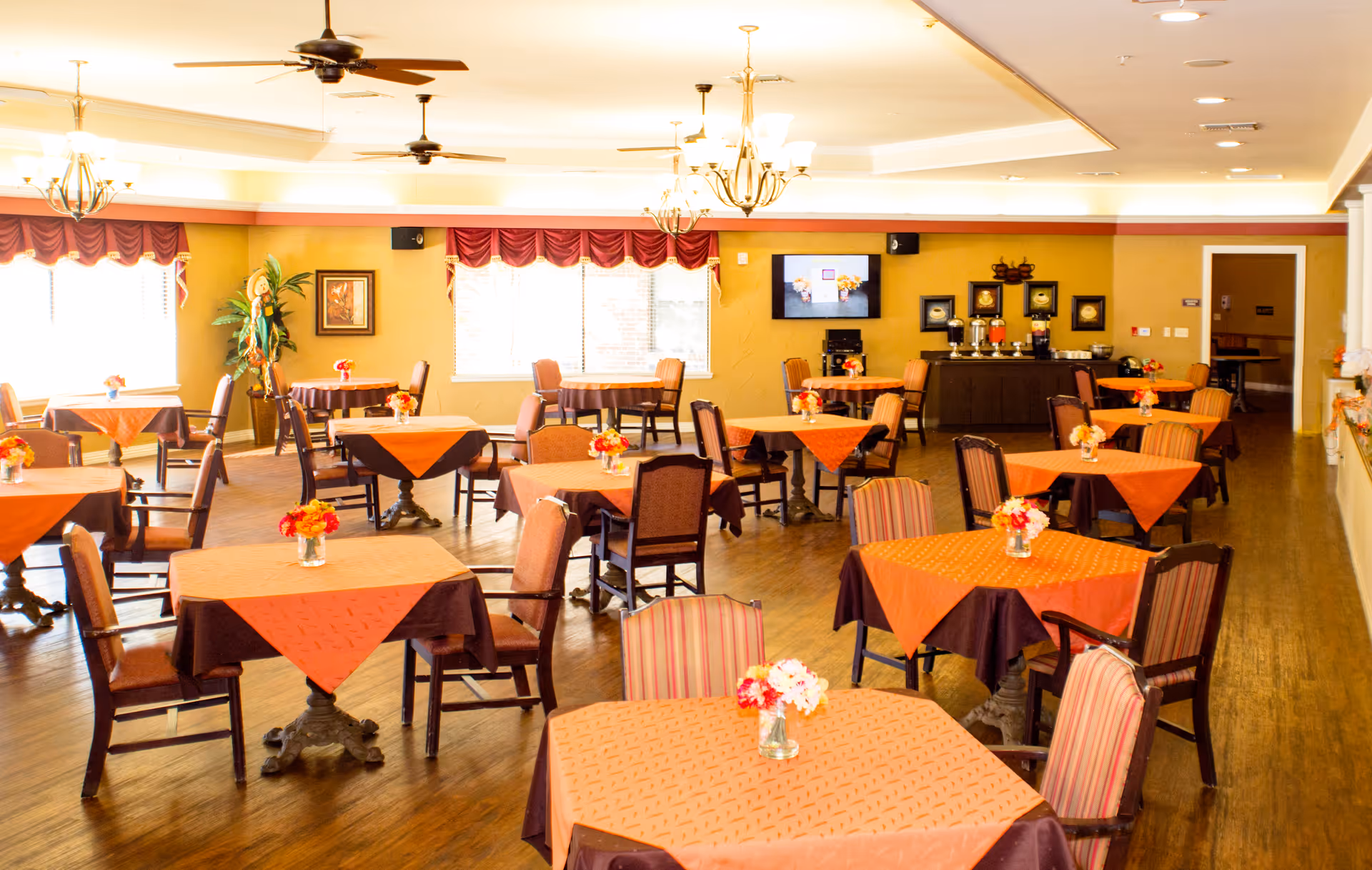 A bright and spacious dining room with multiple square tables covered with orange and brown tablecloths, each adorned with small floral centerpieces. The room features wooden flooring, several chairs around each table, ceiling fans, chandeliers, and large windows with red valances allowing natural light to fill the space. A buffet area with beverage dispensers and a wall-mounted TV is visible in the background.