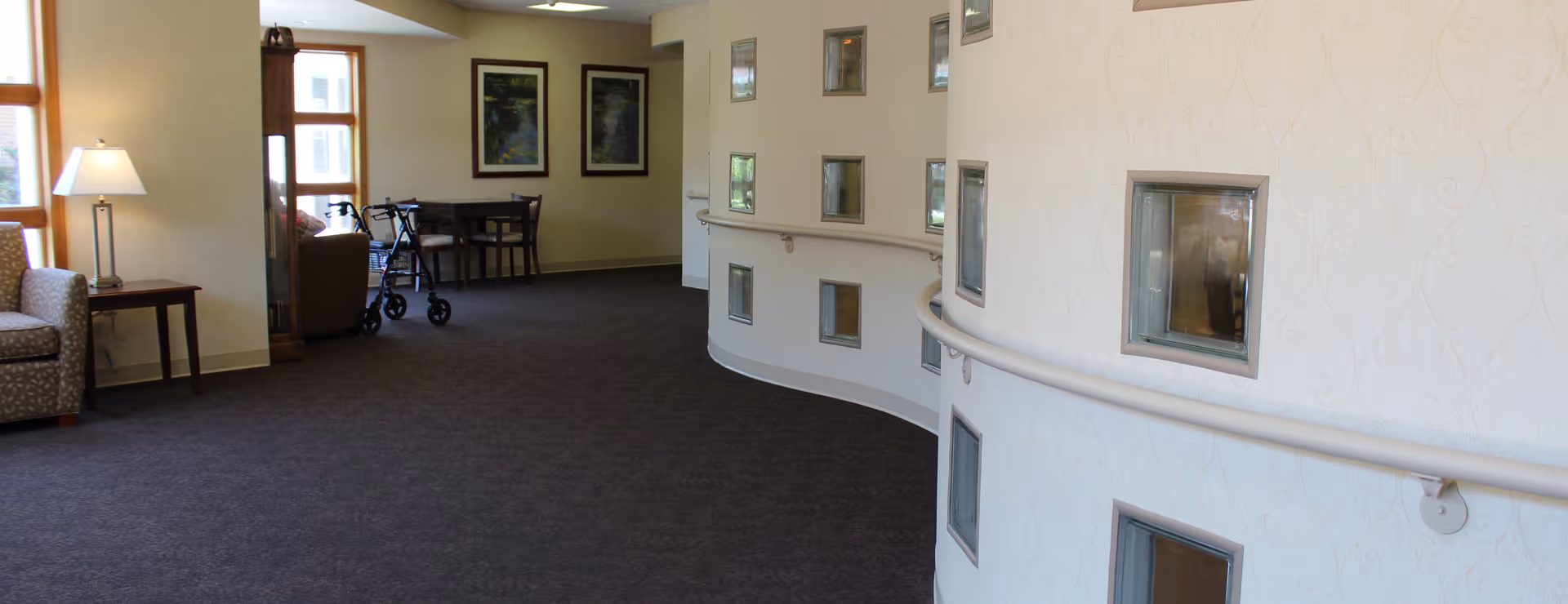 Interior hallway of a senior living facility with a curved wall featuring glass block windows and a handrail. The hallway has carpeted flooring, a small table with a lamp, a patterned armchair, a walker, and a small dining table with chairs near large windows. Two framed paintings hang on the far wall.