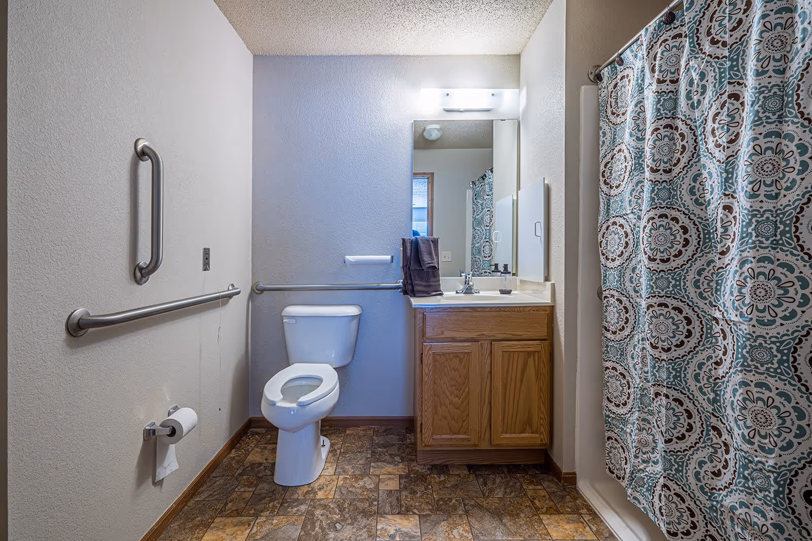 A bathroom with a toilet, a wooden vanity with a sink and mirror above it, a shower with a patterned curtain, and grab bars on the walls. The floor has a tile pattern and there are two dark towels hanging on the vanity.
