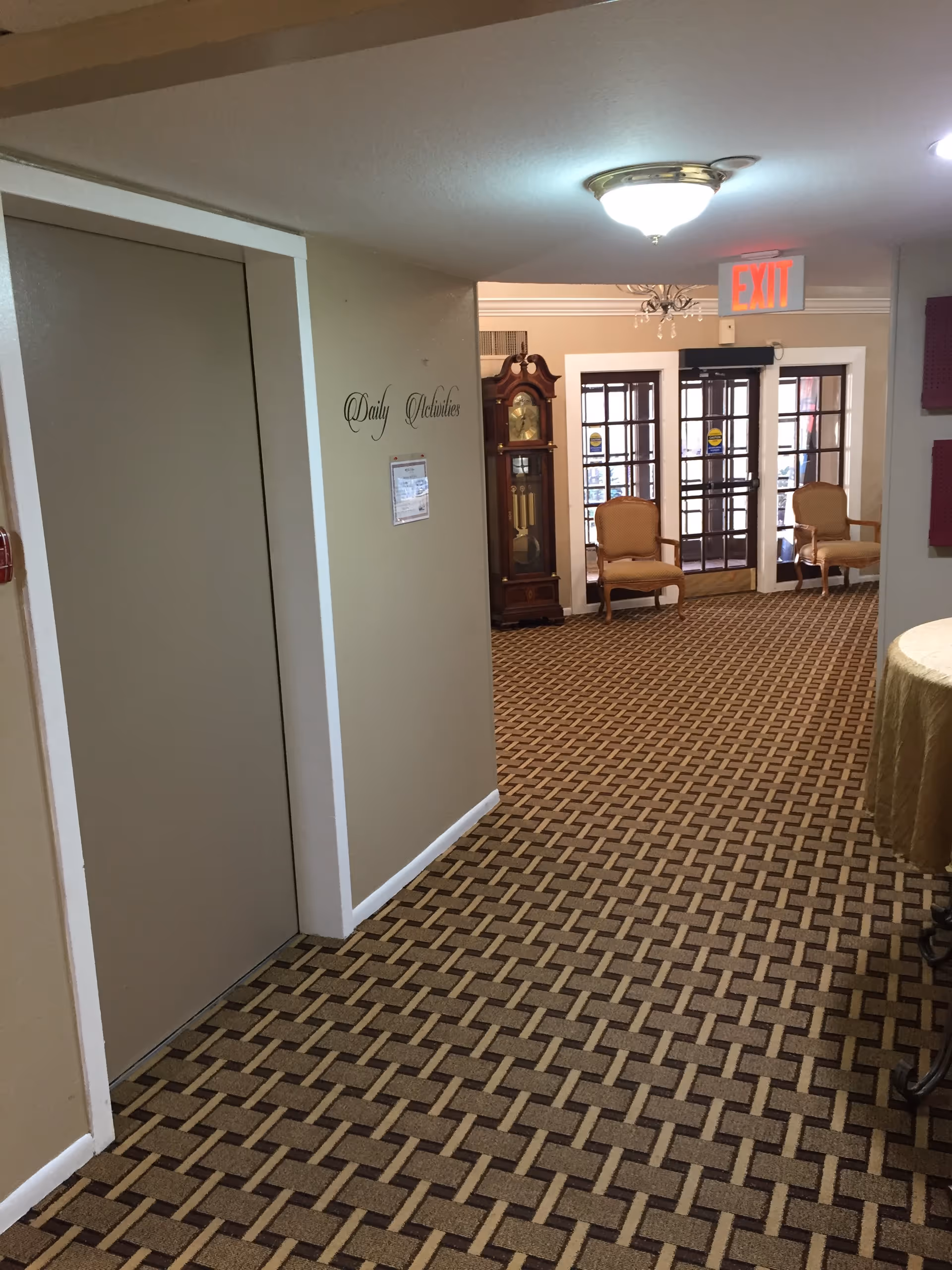 Interior hallway of a senior living facility with patterned carpet, beige walls, and a ceiling light. On the left wall, there is a sign that reads 'Daily Activities' and a closed elevator door. At the end of the hallway, there is a grandfather clock, two upholstered chairs, and glass double doors with an illuminated exit sign above them.