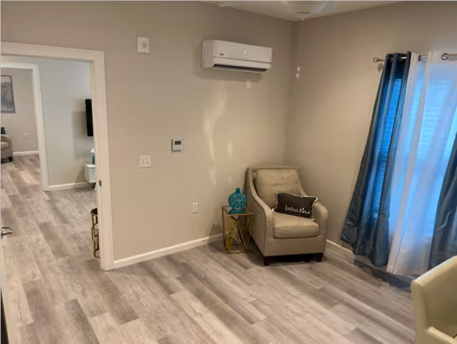Small living room corner with an upholstered chair, decorative side table and pillow, a wall-mounted air-conditioning unit, and a curtained window.
