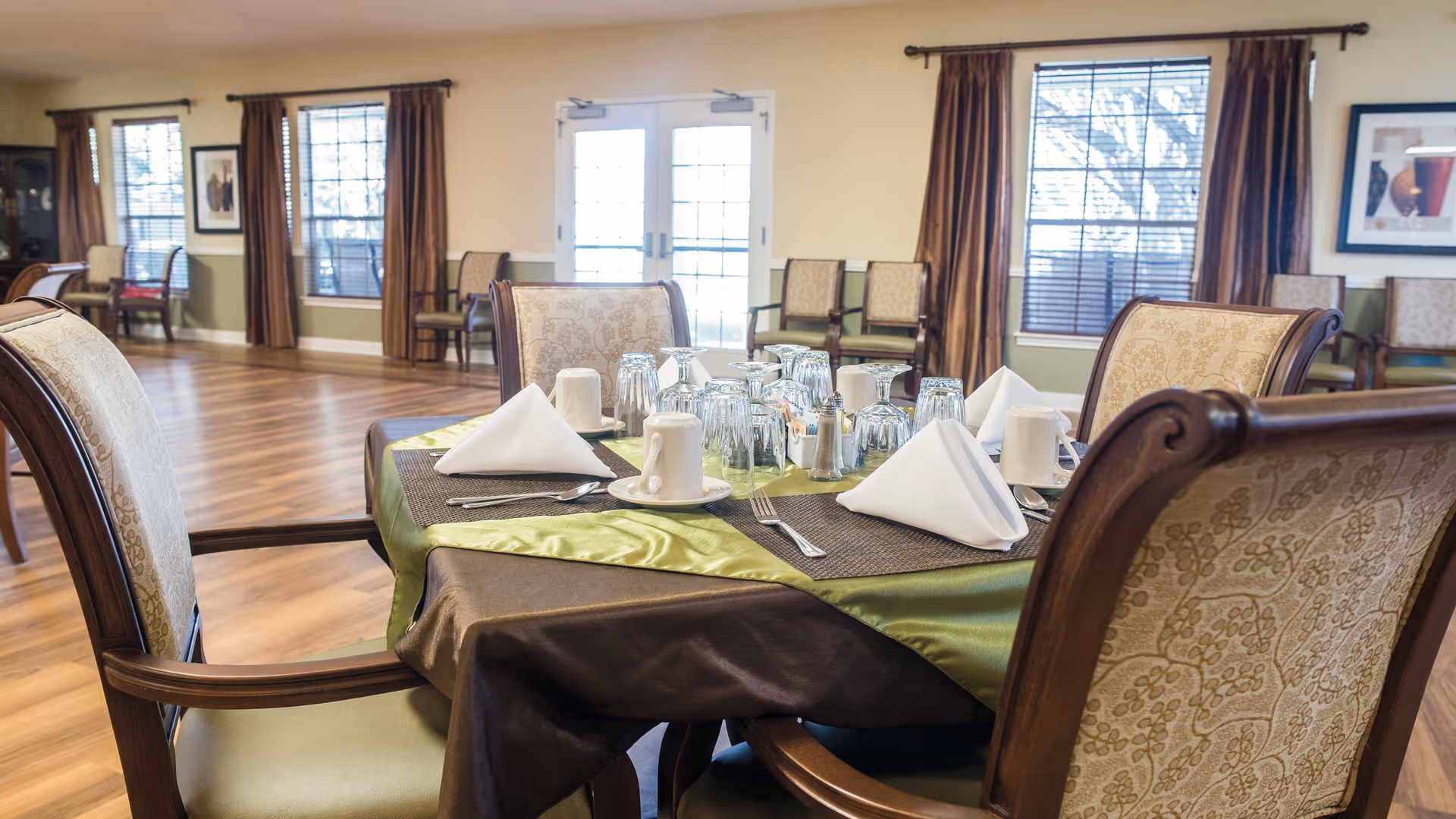 A set dining table with folded napkins, glassware and upholstered chairs in a spacious dining room with wood floors and large windows.