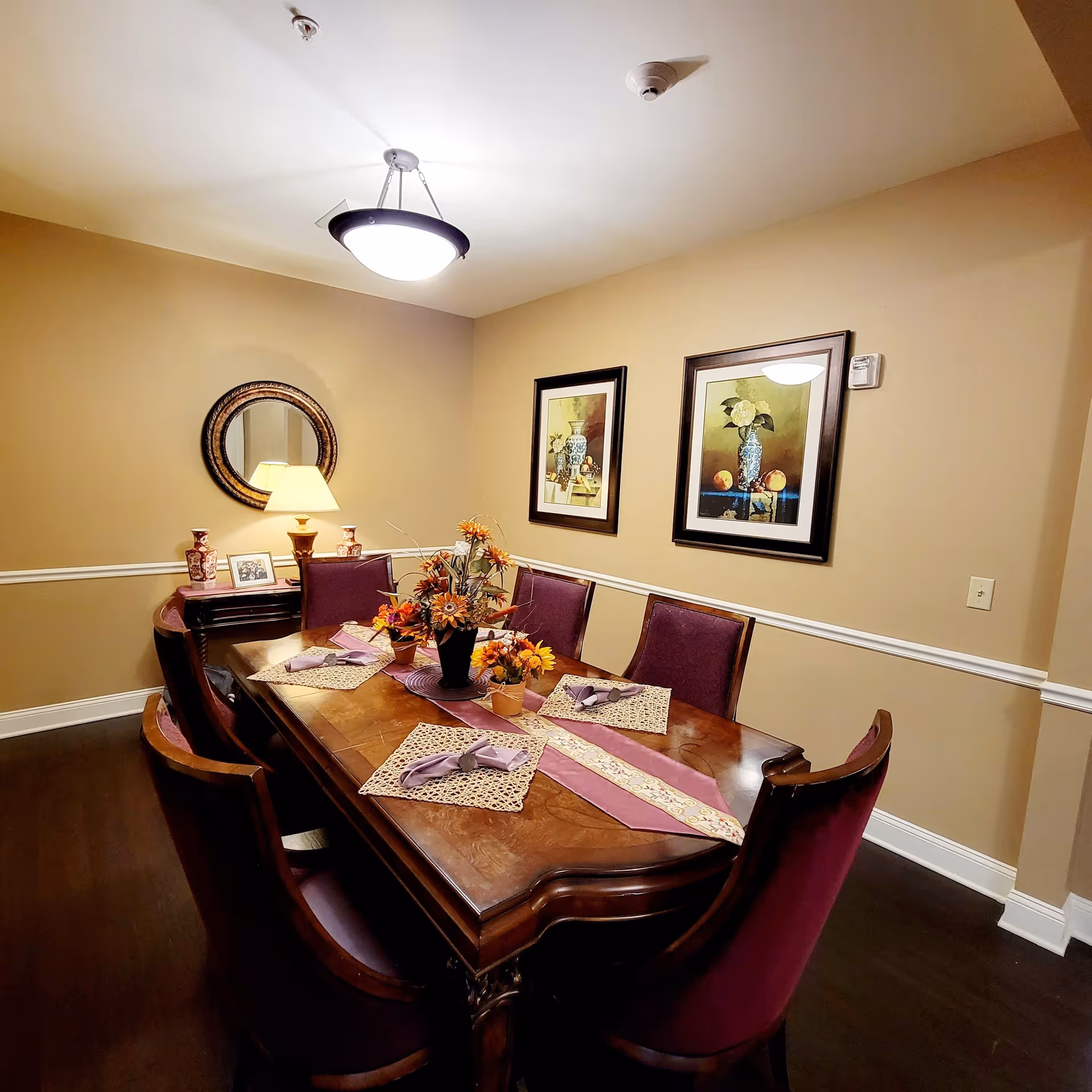 A dining room with a wooden table set for four, featuring purple chairs and floral centerpieces. The walls are beige with white trim, decorated with two framed paintings and a round mirror above a small side table with a lamp and decorative items.