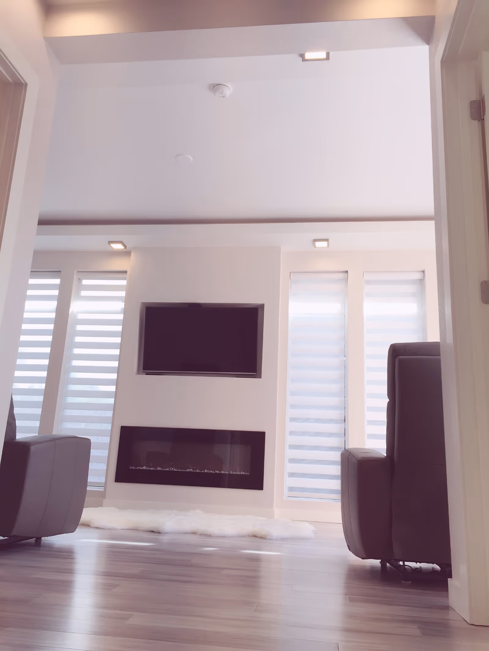 Modern living room with light-colored walls and floor-to-ceiling windows covered by white blinds. A flat-screen TV is mounted above a sleek, rectangular electric fireplace. Two dark-colored armchairs are positioned on either side of the room, and a white fluffy rug lies on the wooden floor in front of the fireplace.