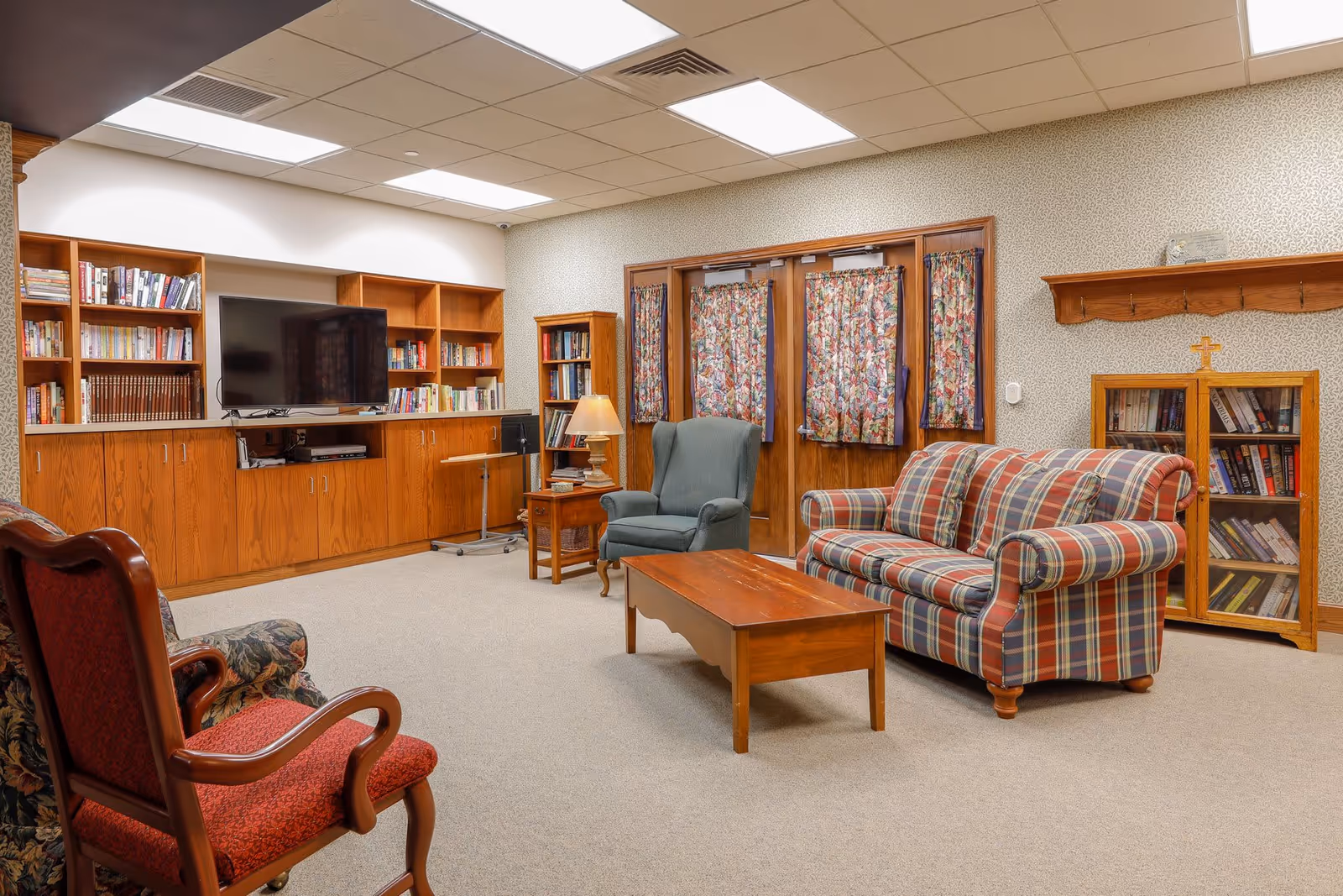 A cozy senior living common room with a plaid sofa, a blue armchair, a wooden coffee table, and two upholstered chairs. The room features built-in wooden bookshelves filled with books, a flat-screen TV, floral curtains on the windows and doors, and a wooden cabinet with a cross on top. The walls have a subtle patterned wallpaper and the ceiling has recessed lighting.
