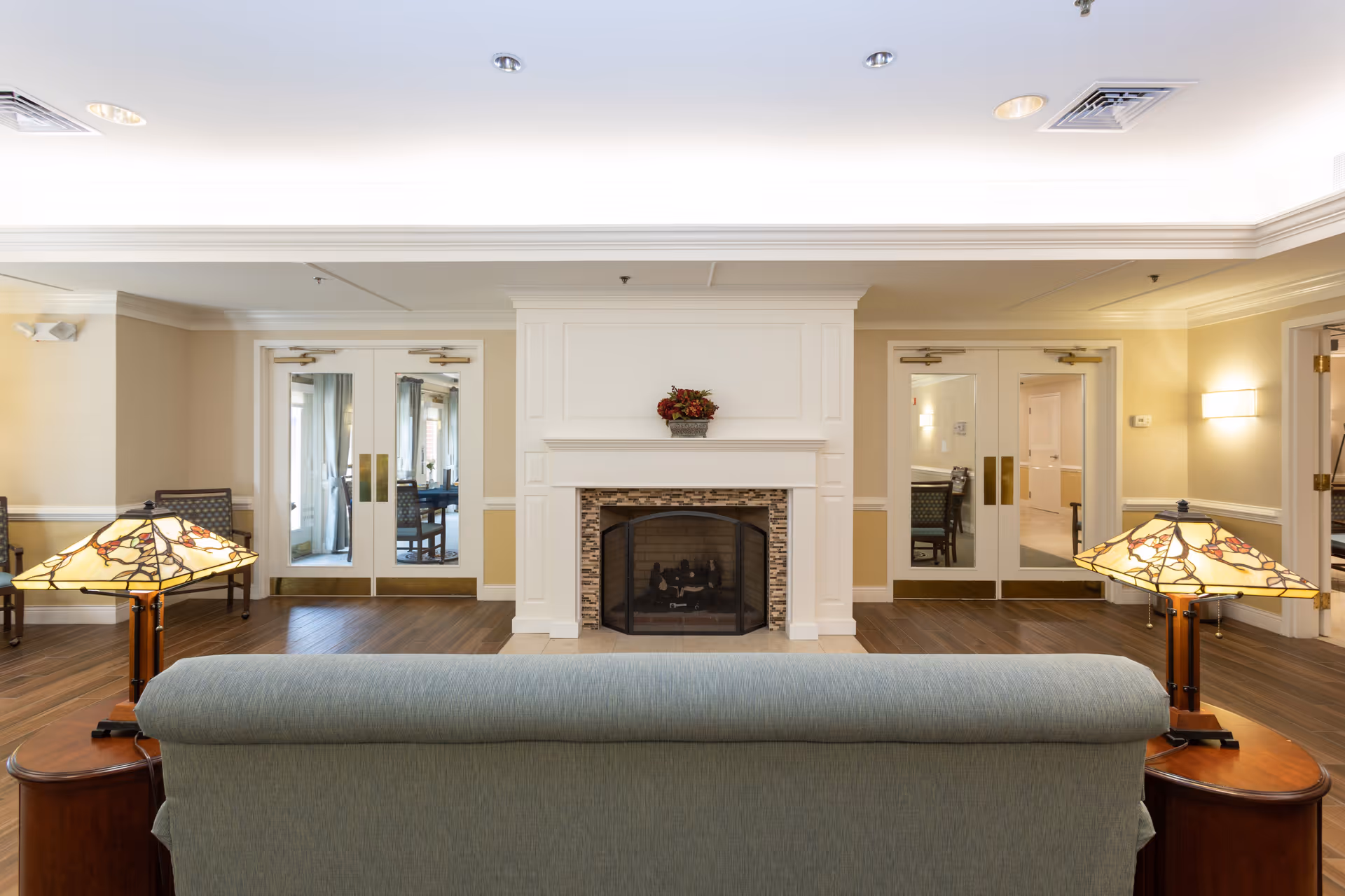A cozy common area with a light blue sofa facing a white fireplace with a decorative plant on the mantel. On either side of the sofa are wooden side tables with Tiffany-style lamps. The room has wooden flooring, beige walls with white trim, and double glass doors on both sides of the fireplace leading to other rooms.