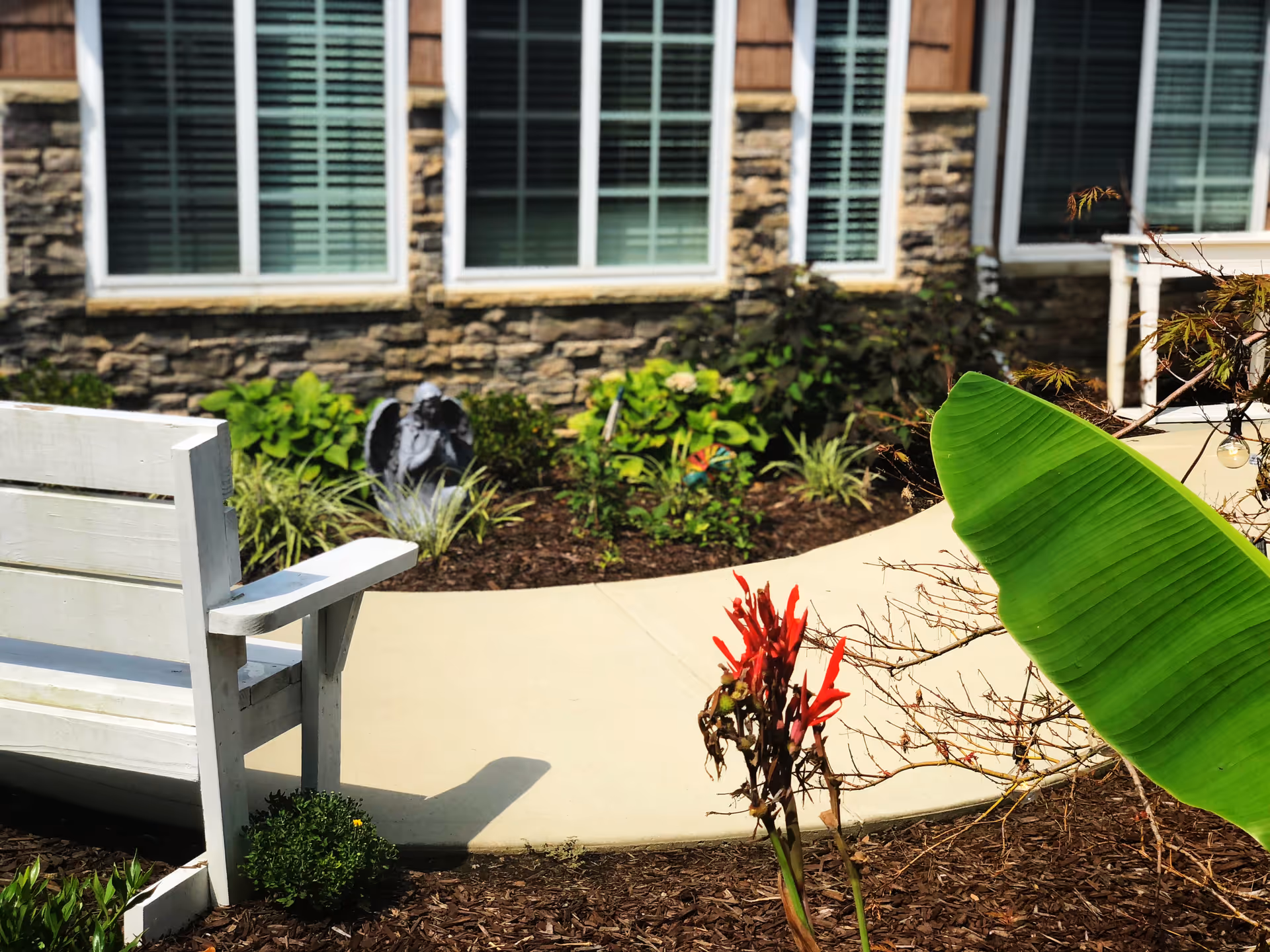 Outdoor garden area with a white wooden bench, a curved concrete pathway, various plants including a large green leaf and red flowers, and a stone and wood building facade with windows in the background.