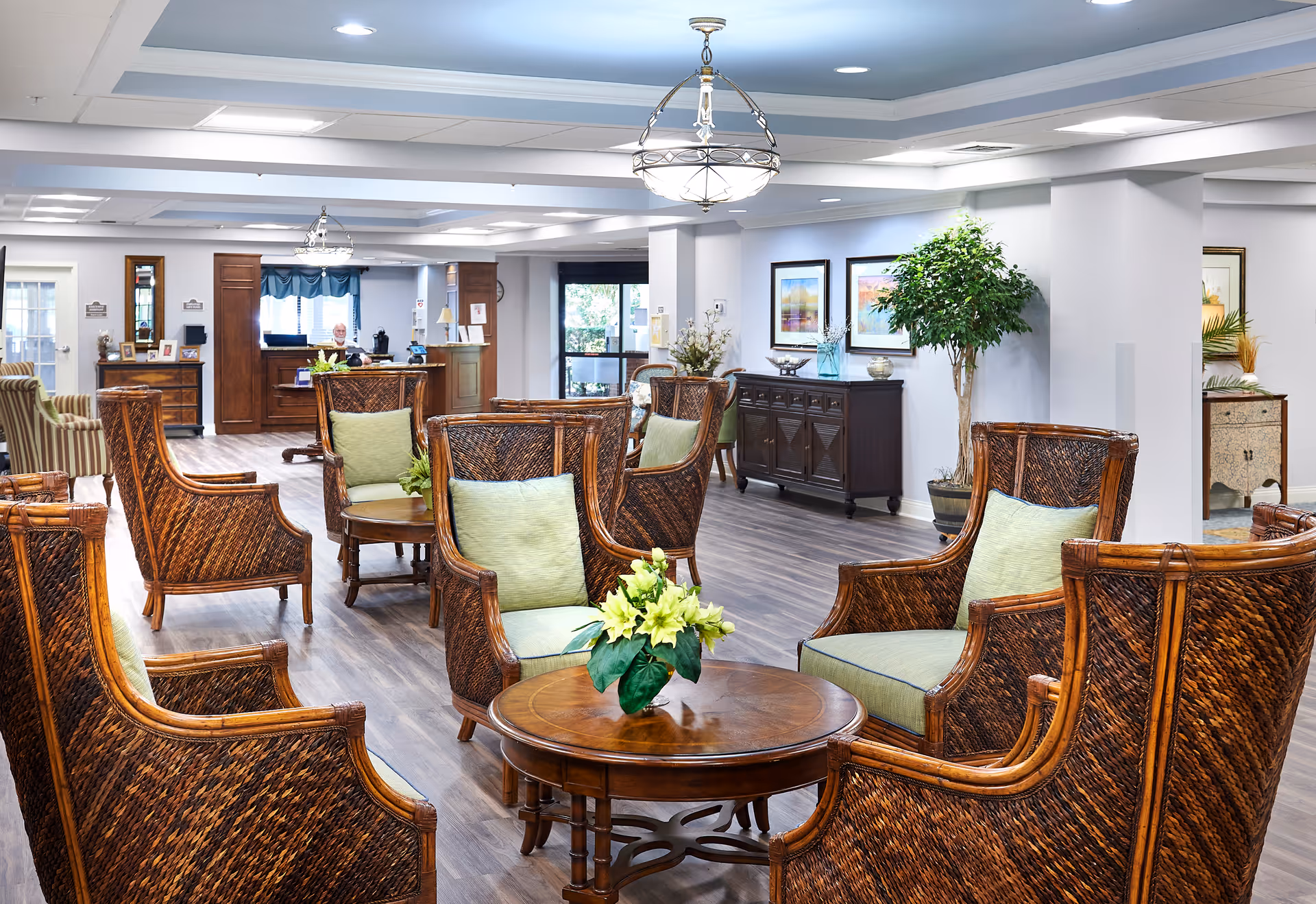 Bright furnished senior living lounge with wicker armchairs around round tables, decorative plants, and a reception desk in the background.