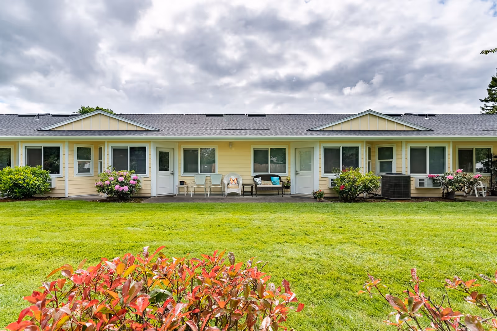 Exterior view of a single-story yellow assisted living facility building with multiple windows and doors, a well-maintained green lawn in front, flowering bushes, and a cloudy sky overhead.