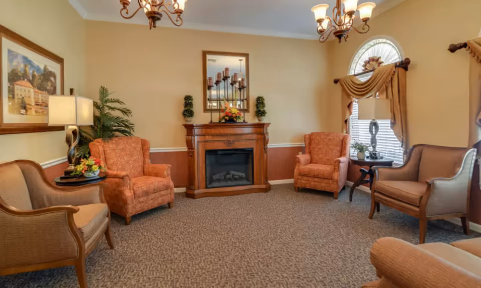 Cozy sitting room with upholstered armchairs arranged around a decorative fireplace under chandeliers.