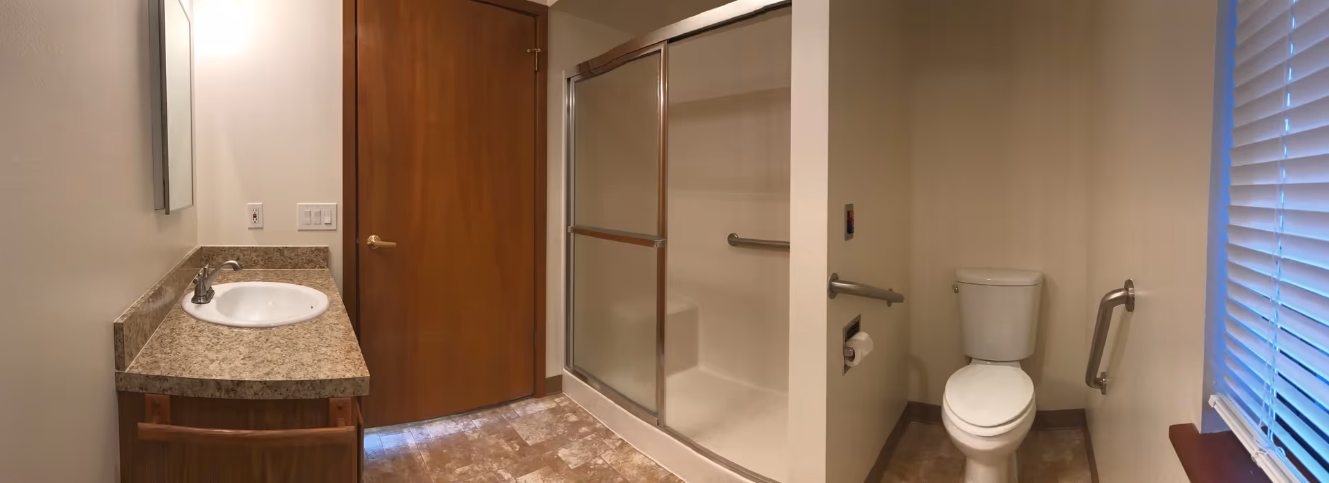 A bathroom with a single sink and countertop on the left, a wooden door in the center, a shower with sliding glass doors next to the door, and a toilet with grab bars on the right. There is a window with blinds on the far right side.