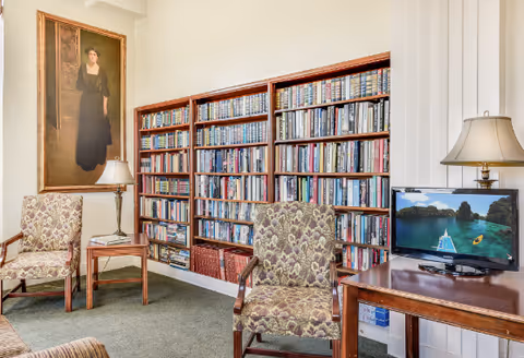 A cozy interior room with two patterned armchairs, a wooden side table with a lamp, a large bookshelf filled with books, a flat-screen TV on a wooden table displaying a scenic water and mountain view, and a large portrait painting on the wall.