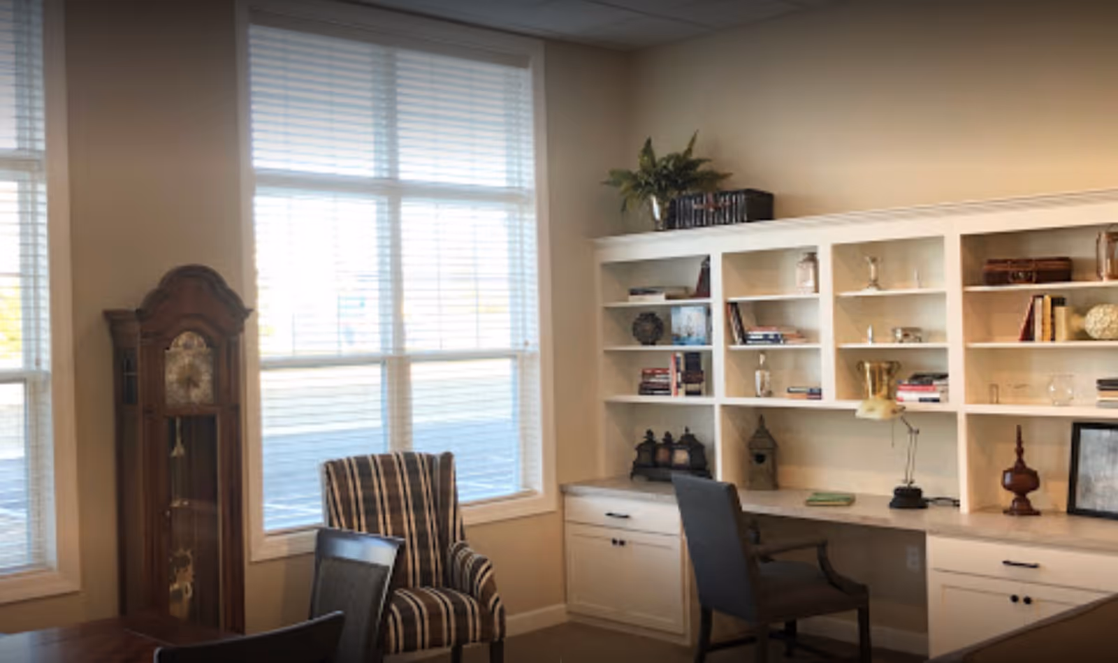 A cozy interior room with large windows covered by white blinds, a tall wooden grandfather clock, a striped armchair, a desk with a chair, and built-in white shelves filled with books and decorative items.