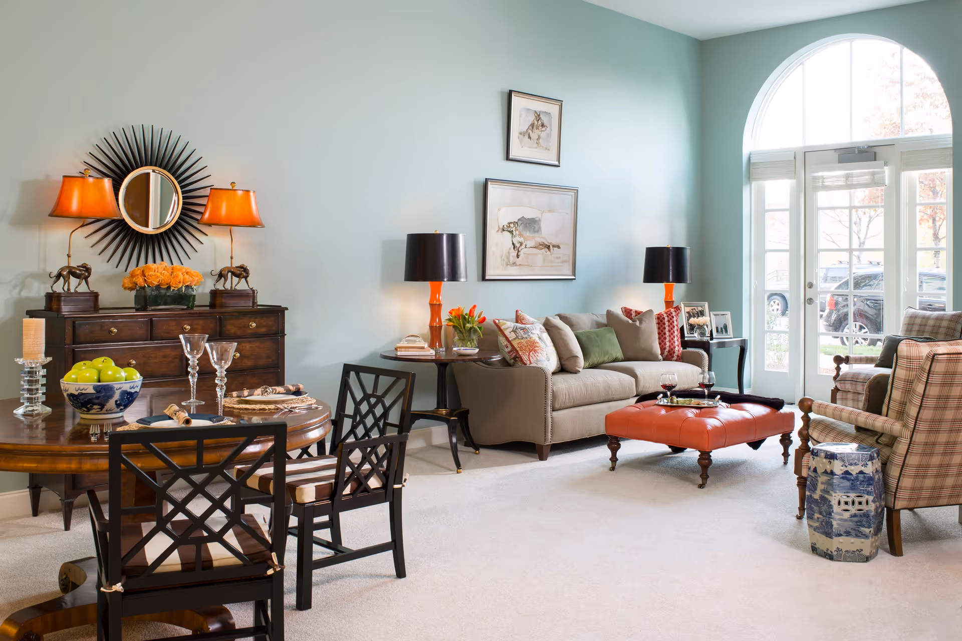 A bright and elegant living room with a beige sofa adorned with multiple cushions, two plaid armchairs, and a red leather ottoman serving as a coffee table. The room features a round wooden dining table with four chairs, a dark wooden sideboard with two orange lamps and decorative items, and large arched windows letting in natural light. The walls are painted light blue and decorated with framed artwork.