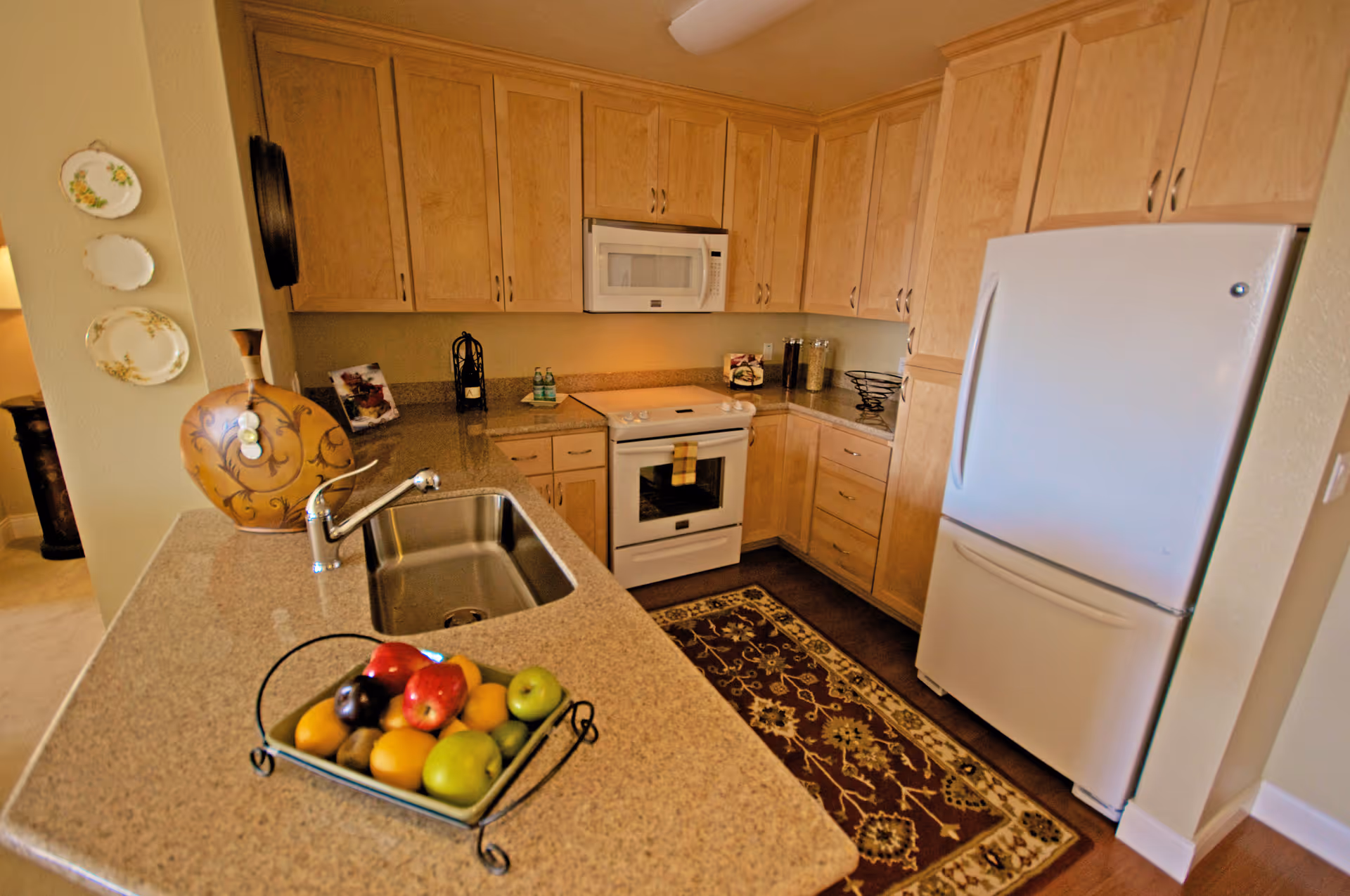 A kitchen with light wood cabinets, a white refrigerator, a white stove with an oven, a white microwave above the stove, and a granite countertop with a built-in sink. On the countertop is a decorative tray with various fruits. There is a decorative rug on the wooden floor in front of the stove. Plates are hung on the wall to the left, and a large decorative vase is placed on the counter near the wall.