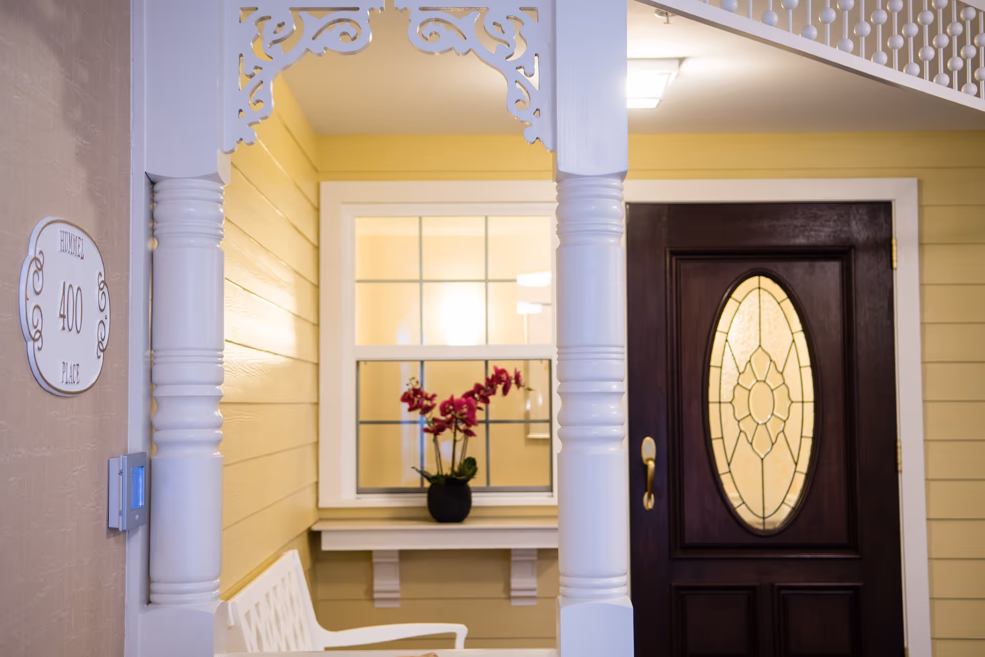 Entrance area of a senior living facility with a dark wooden door featuring an oval stained glass window, yellow walls, a white decorative column and trim, a white bench, and a window with a shelf holding a potted plant with red flowers.