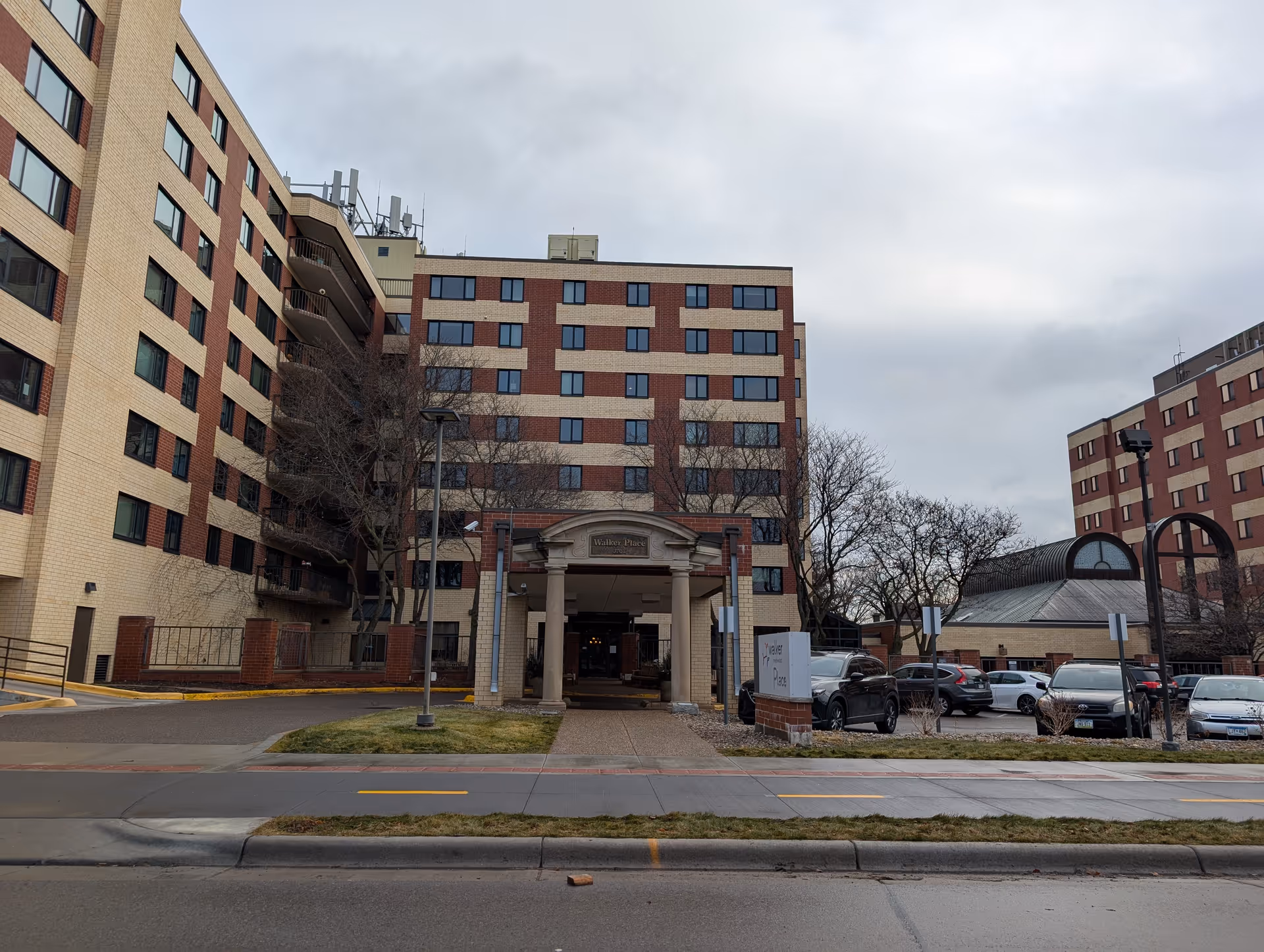Exterior view of a multi-story senior living facility named Walker Place by Vivie, showing the main entrance with columns and a covered driveway. Several cars are parked in the parking lot to the right, and leafless trees are visible around the building under a cloudy sky.