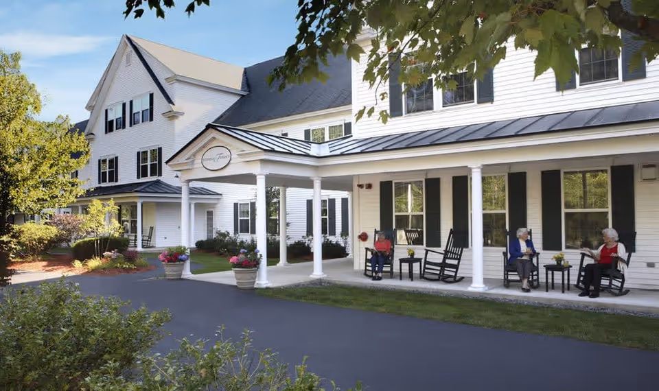 Exterior view of a white senior living facility building with a covered porch. Three elderly women are sitting on rocking chairs on the porch, engaged in conversation. The area is surrounded by greenery and potted flowers, with a clear blue sky overhead.