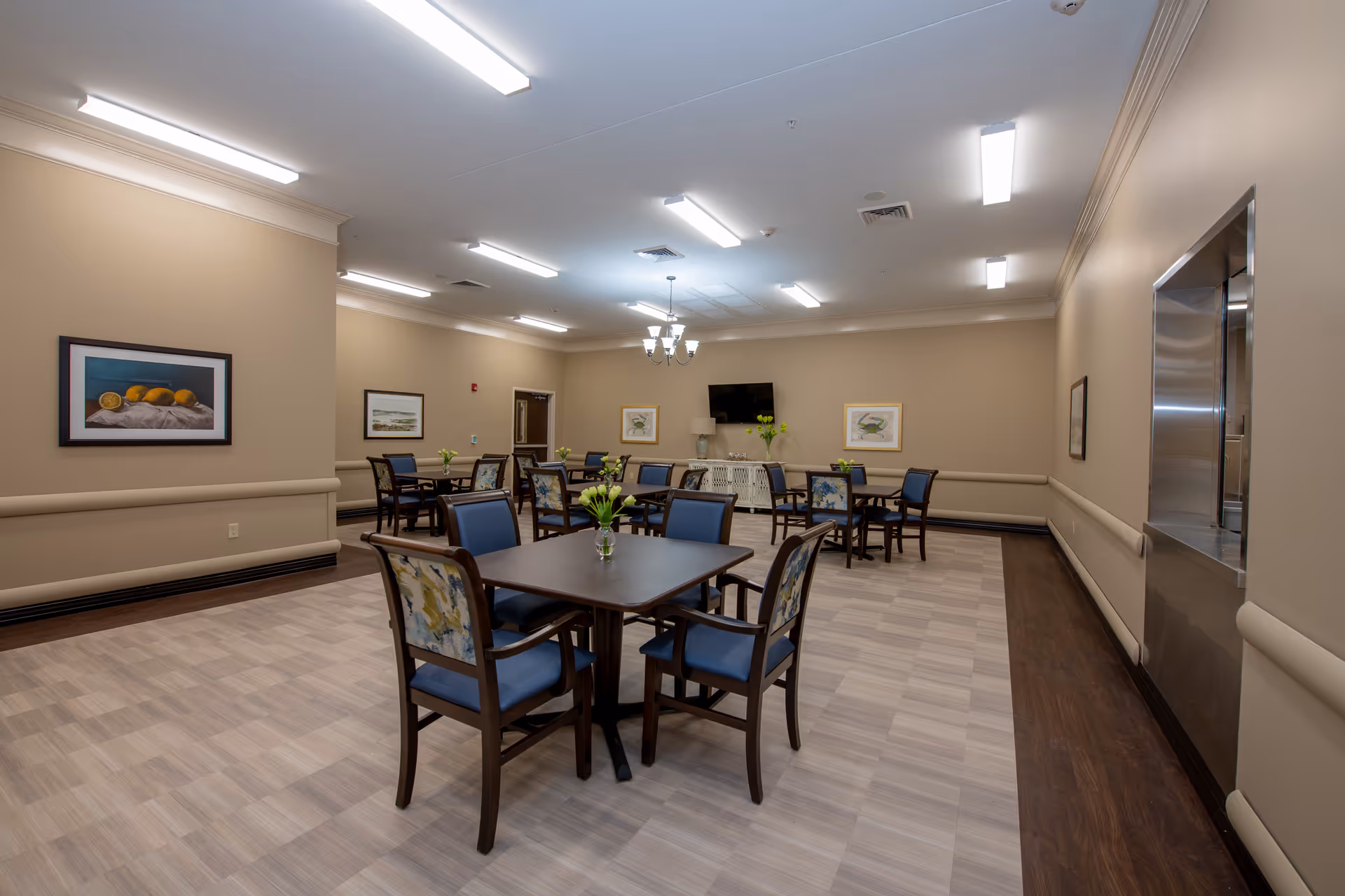 A spacious dining room in a senior living facility with multiple square tables, each surrounded by four chairs with blue cushions and floral-patterned backs. The room has beige walls adorned with framed artwork, a flat-screen TV mounted on the far wall, and a chandelier hanging from the ceiling. The floor features a combination of light and dark wood tones, and there is a serving window on the right wall.