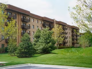 Exterior view of a multi-story apartment building with balconies, surrounded by green trees and a well-maintained lawn under a partly cloudy sky.