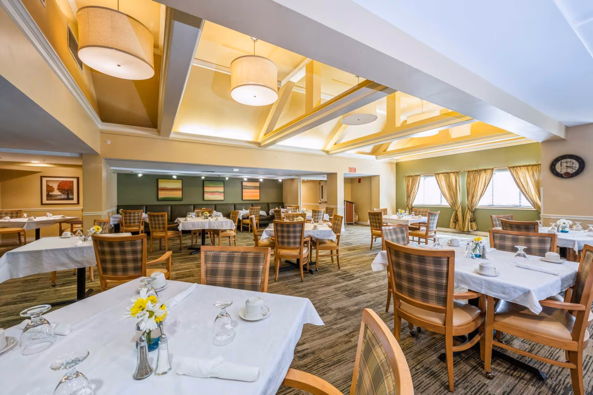 A bright and spacious dining room with multiple tables covered in white tablecloths, each set with cups, glasses, napkins, and small flower arrangements. The room features plaid cushioned chairs, large windows with gold curtains, and a ceiling with recessed lighting and hanging lamps.