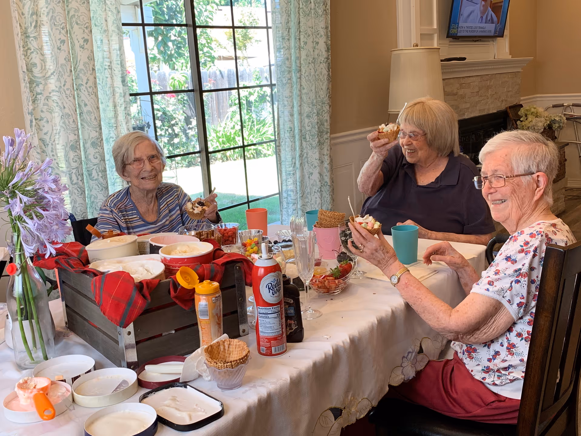 Three elderly women sitting around a dining table enjoying ice cream sundaes. The table is covered with a white tablecloth and has various ice cream toppings, cones, and whipped cream. There is a large window with patterned curtains letting in natural light, and a vase with purple flowers is on the table. The room has a cozy atmosphere with a fireplace and a TV mounted on the wall in the background.