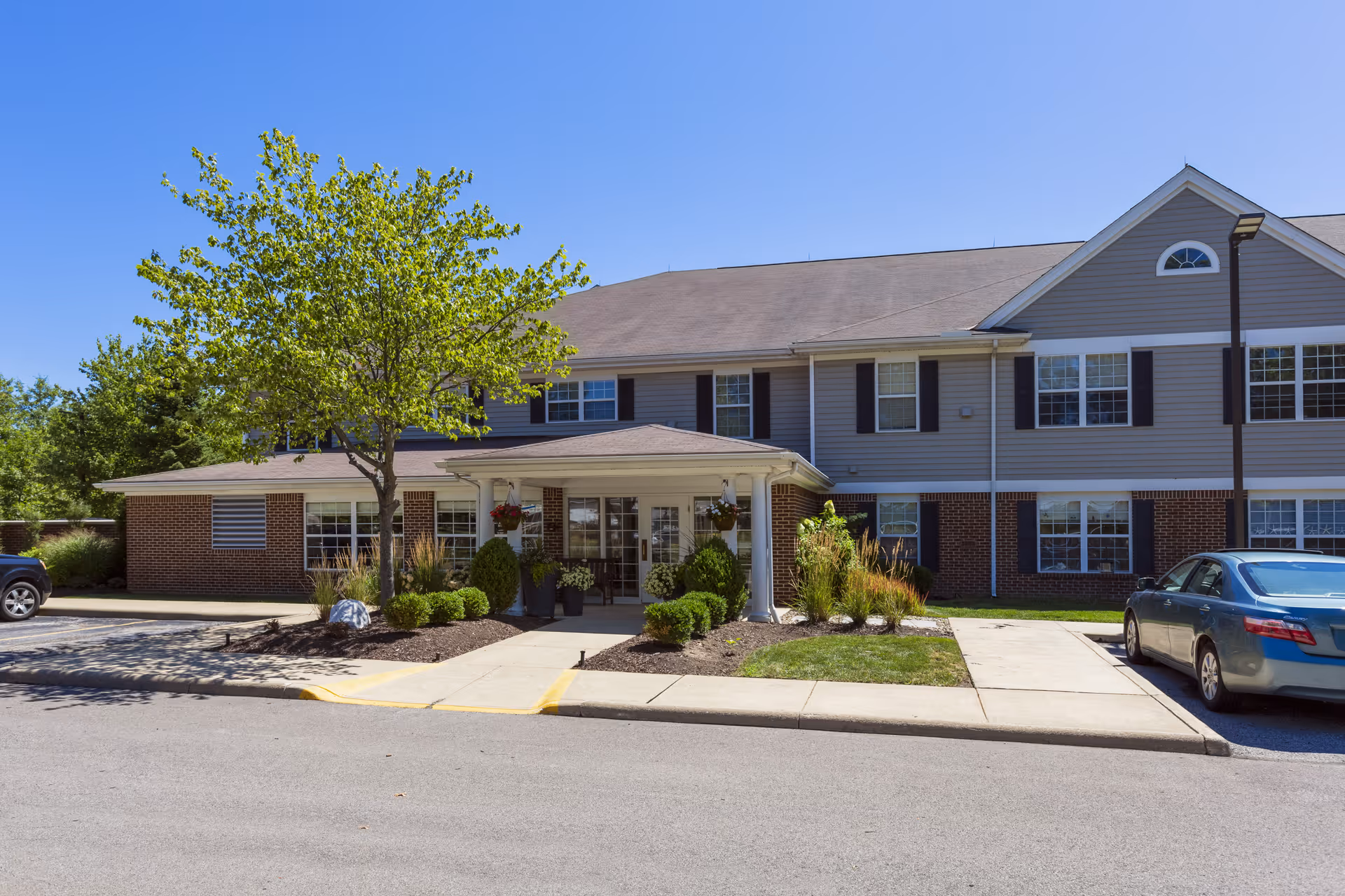 Exterior view of Independence Village of Avon Lake, showing a two-story building with a covered entrance, landscaped greenery, and parked cars under a clear blue sky.