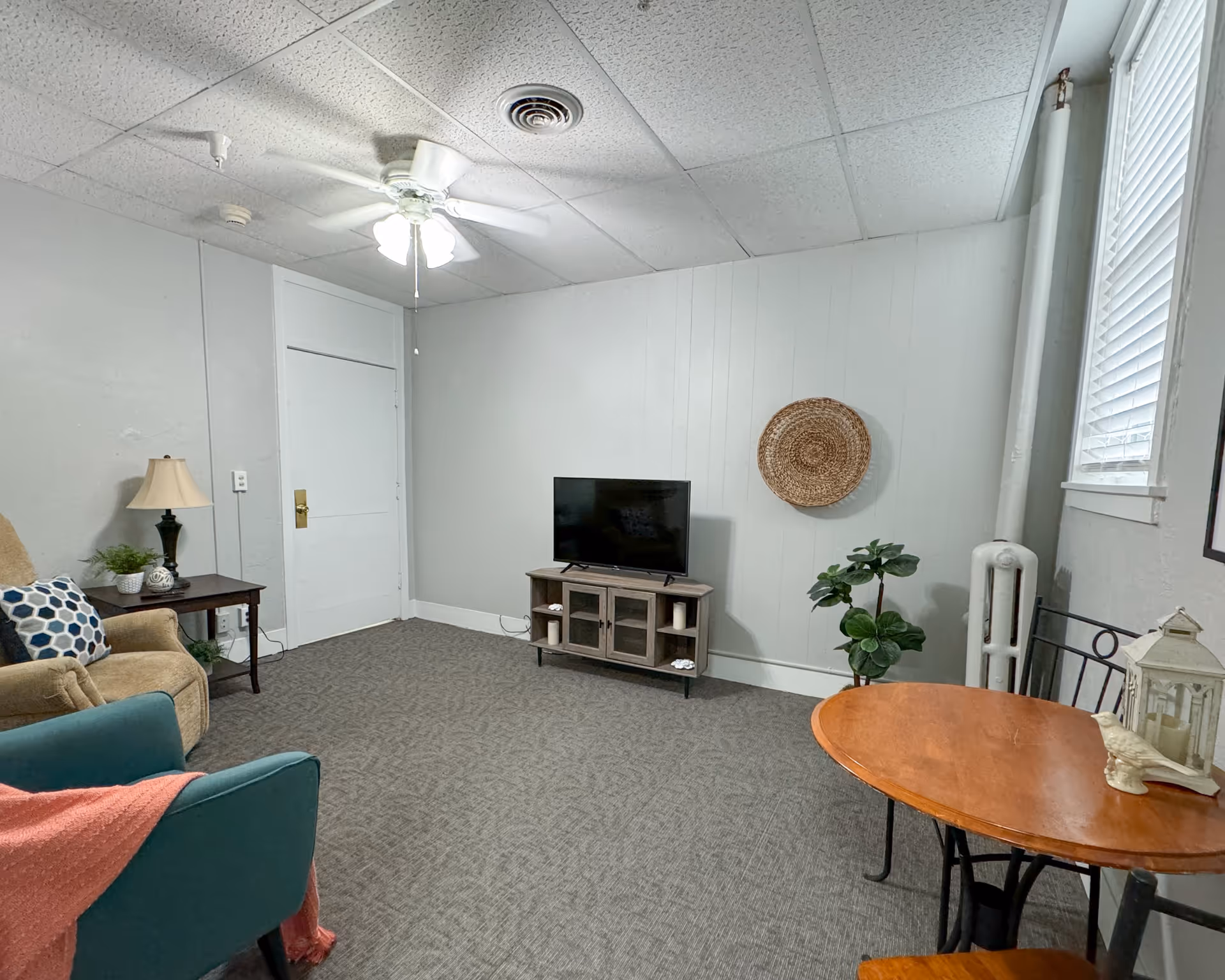 A small living room with a beige armchair and a teal chair with a pink throw blanket. There is a wooden side table with a lamp and plant next to the armchair. A flat-screen TV sits on a wooden TV stand against a white wall with a woven circular wall decoration above it. A small round wooden table with two chairs is positioned near a window with blinds. The room has a ceiling fan with lights and a carpeted floor.