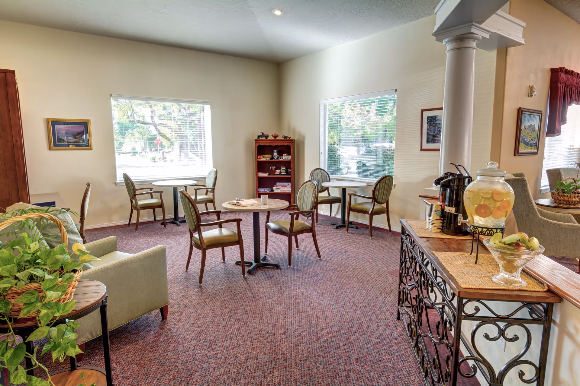 A cozy senior living common area with several small round tables and chairs arranged near two large windows. There is a green armchair with a plant on a small table beside it. A wooden shelf with books and games is against the wall. A decorative wrought iron and wood console table holds a glass dispenser with fruit-infused water, coffee urns, and a bowl of bananas. The room is carpeted and has light-colored walls with framed artwork.