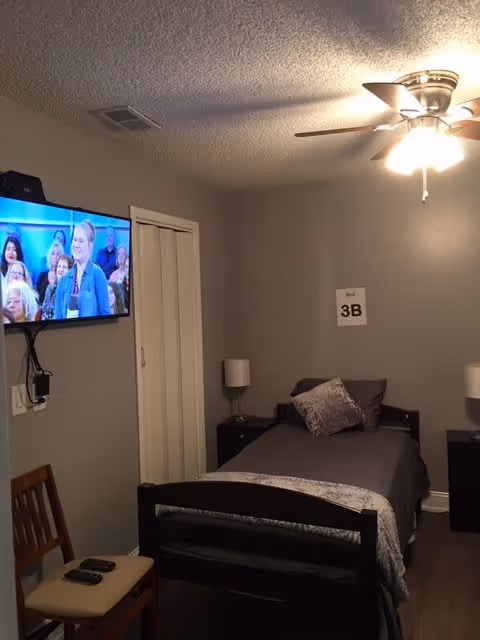 A small bedroom with a single bed made with gray bedding and a decorative pillow. There are two bedside tables with lamps on either side of the bed. A ceiling fan with lights is on the ceiling. A flat-screen TV is mounted on the wall showing a woman speaking in front of an audience. A wooden chair with two remote controls on the seat is positioned near the bed. A sign on the wall above the bed reads 'Room 3B'.