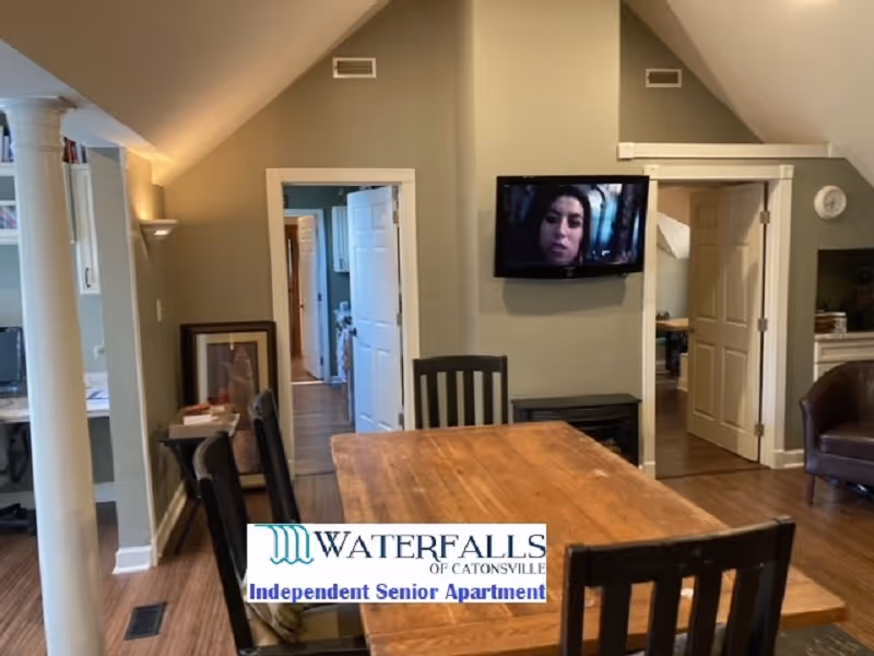 Interior view of a senior living facility room with a wooden dining table and chairs in the center. The room has beige walls and a sloped ceiling. A flat-screen TV is mounted on the wall showing a woman's face. There are two open doorways leading to other rooms, and a brown armchair is visible on the right side. The floor is wooden, and there is a white column on the left side near a small desk area.