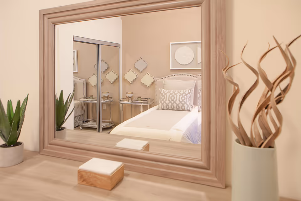 Reflection in a large wooden-framed mirror showing a neatly made bed with decorative pillows, a small round side table with candles, and a closet with sliding mirrored doors. On the dresser in front of the mirror are a small potted plant and a vase with dried decorative branches.