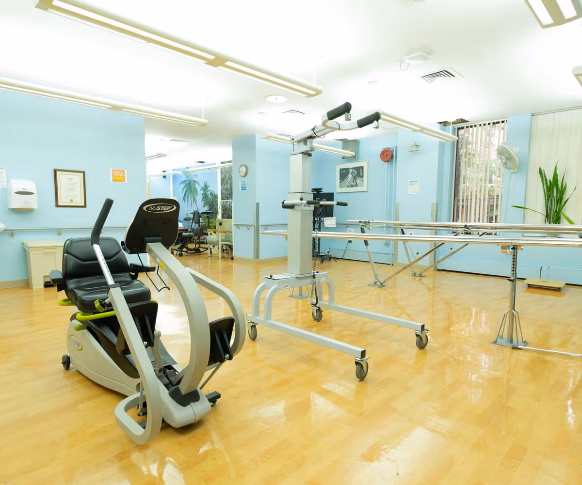 Bright rehabilitation/therapy room with exercise equipment, parallel bars, and a recumbent bike on a polished wood floor.