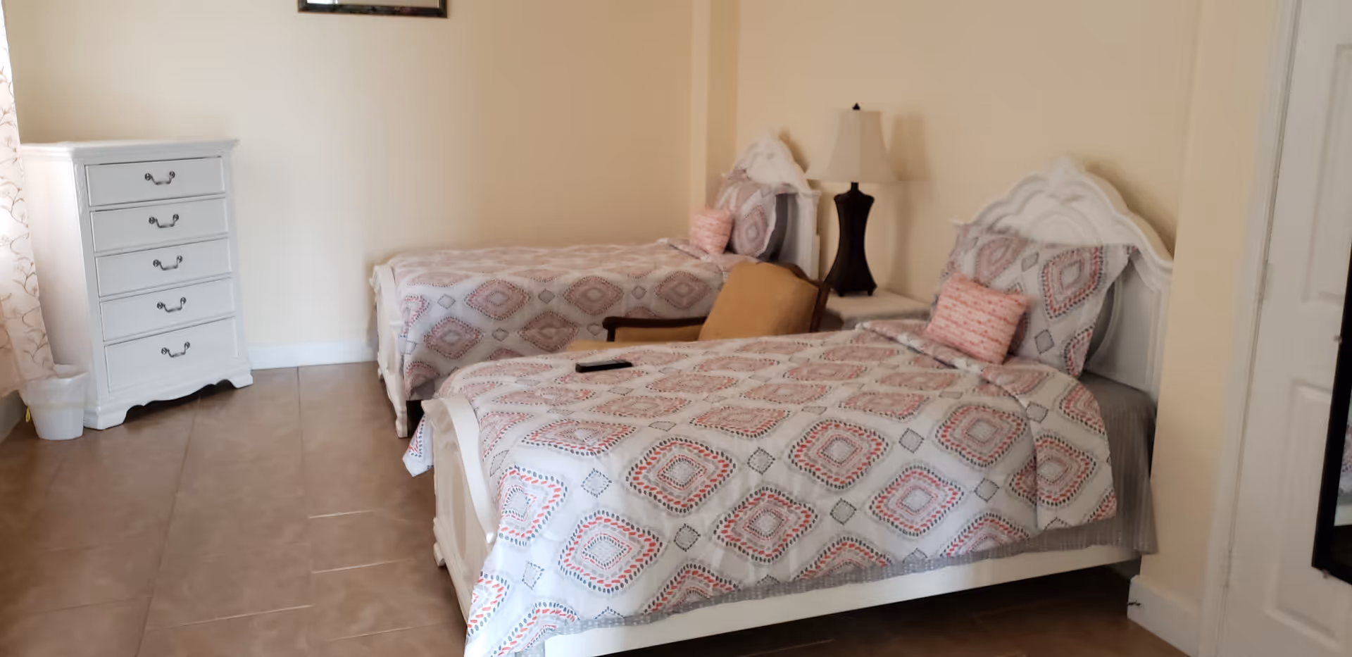 A bright bedroom with two twin beds featuring patterned bedspreads, a white dresser, and a bedside lamp.