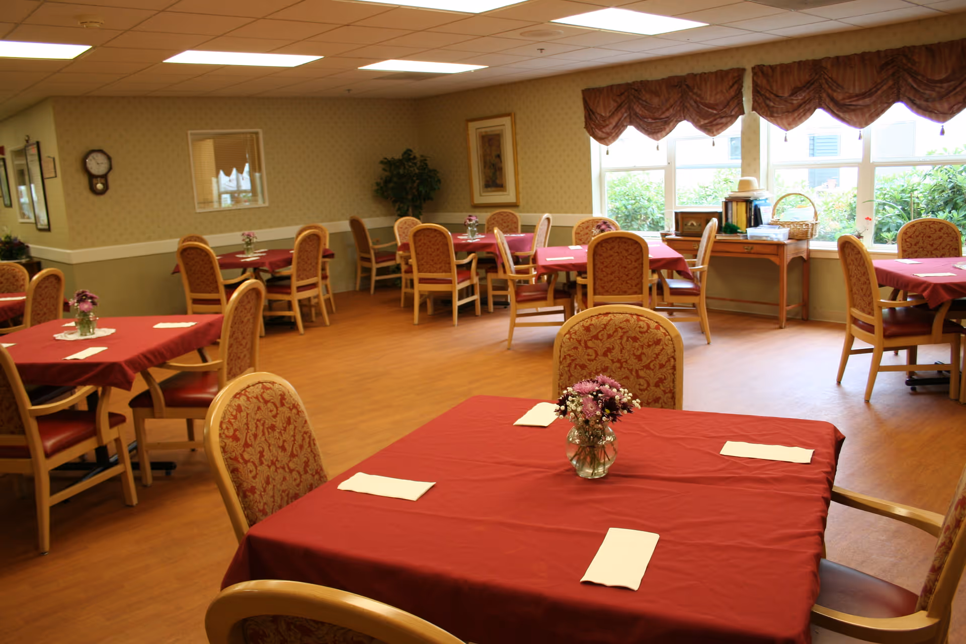 Dining room with multiple tables covered in red tablecloths, floral centerpieces, and wooden chairs by large windows.