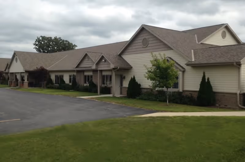 Single-story beige assisted living building with pitched roofs, small landscaped front area, and an adjacent parking lot under a cloudy sky.