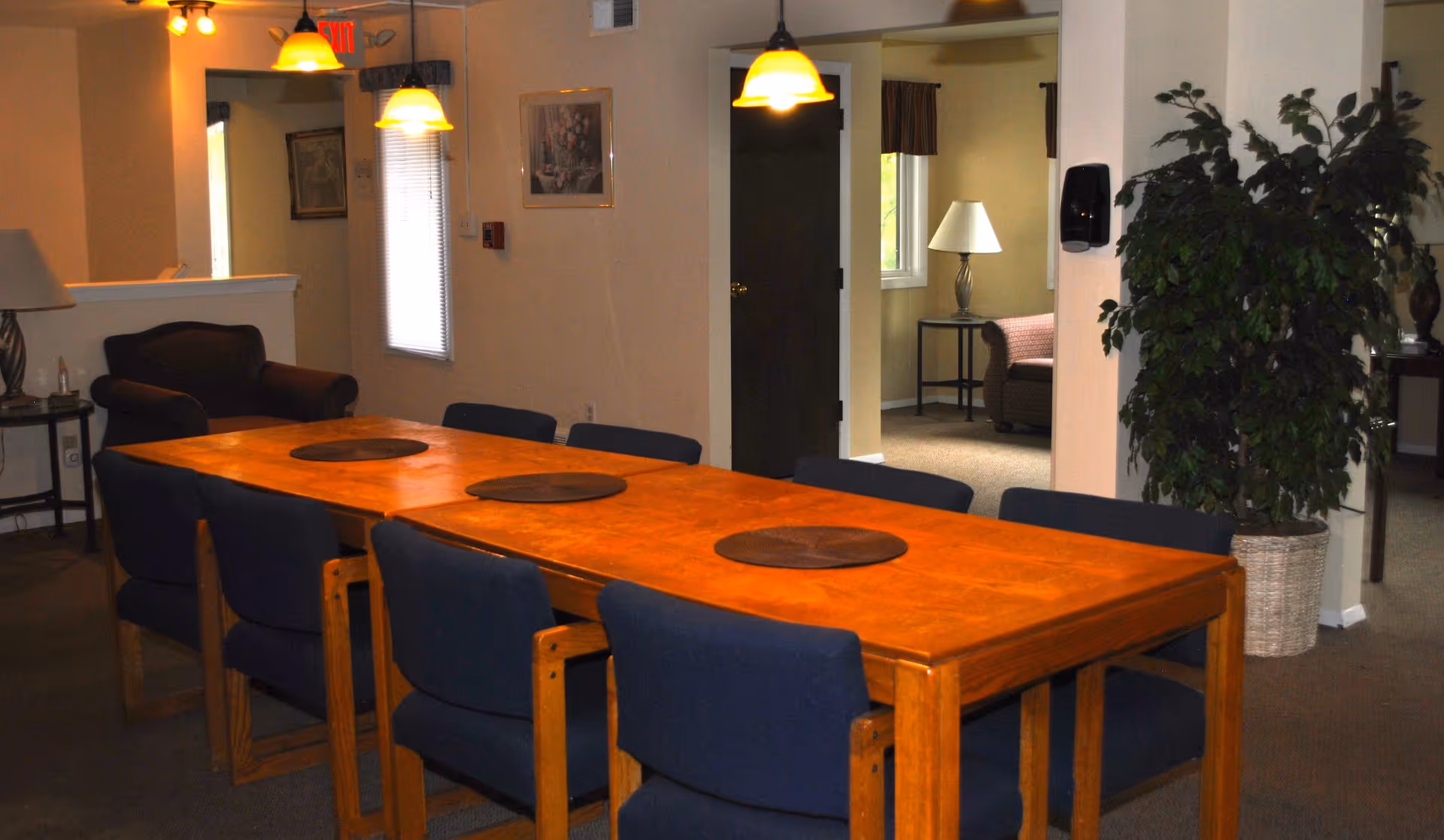 A dining room with a long wooden table surrounded by eight blue cushioned chairs. The table has three round placemats. The room is softly lit by three hanging pendant lights. In the background, there is a small sitting area with an armchair, a side table with a lamp, a large potted plant, and a window with curtains.