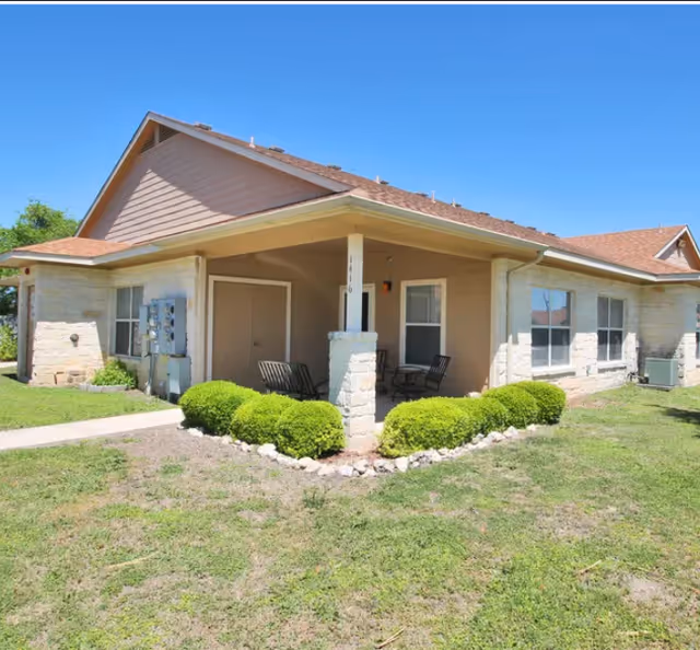 Single-story beige senior living building with a covered porch, shrubs, and seating under a blue sky.