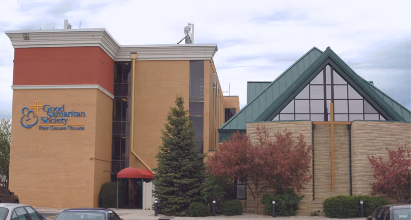 Front exterior of the Good Samaritan Society - Fort Collins Village building showing the facility sign, a peaked glass entry with a cross, and surrounding landscaping.