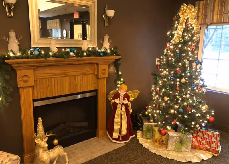 A cozy interior corner with a wooden fireplace mantel and a decorated, lit Christmas tree surrounded by wrapped presents and holiday figurines.