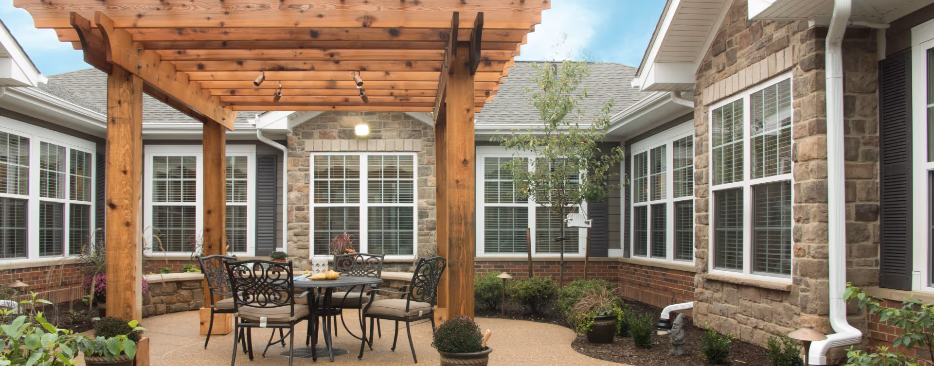 Outdoor courtyard area at Bickford of Spotsylvania featuring a wooden pergola, a round metal table with four cushioned chairs, surrounded by stone and brick building walls with multiple windows and some landscaping including small trees and plants.