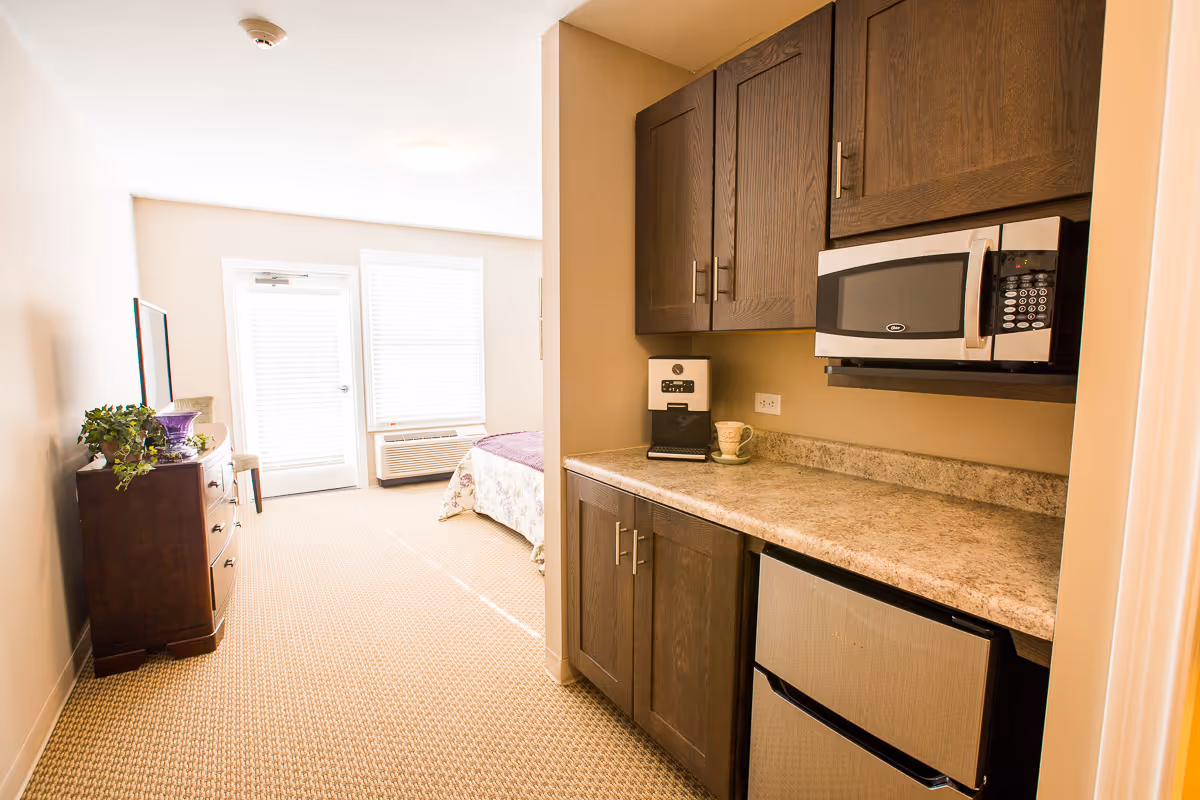 Interior view of a senior living facility room showing a small kitchenette with dark wood cabinets, a microwave, a coffee maker, and a mini refrigerator. The room extends into a bedroom area with a bed, a dresser with a plant and decorative items, a chair, and a door with a window covered by blinds letting in natural light.
