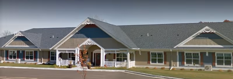 Exterior view of a single-story senior care facility building with a gray roof, beige siding, white trim, and decorative gable accents. The building has multiple windows and a covered entrance with white railings. There is a paved driveway and some landscaping with grass and small plants in front.
