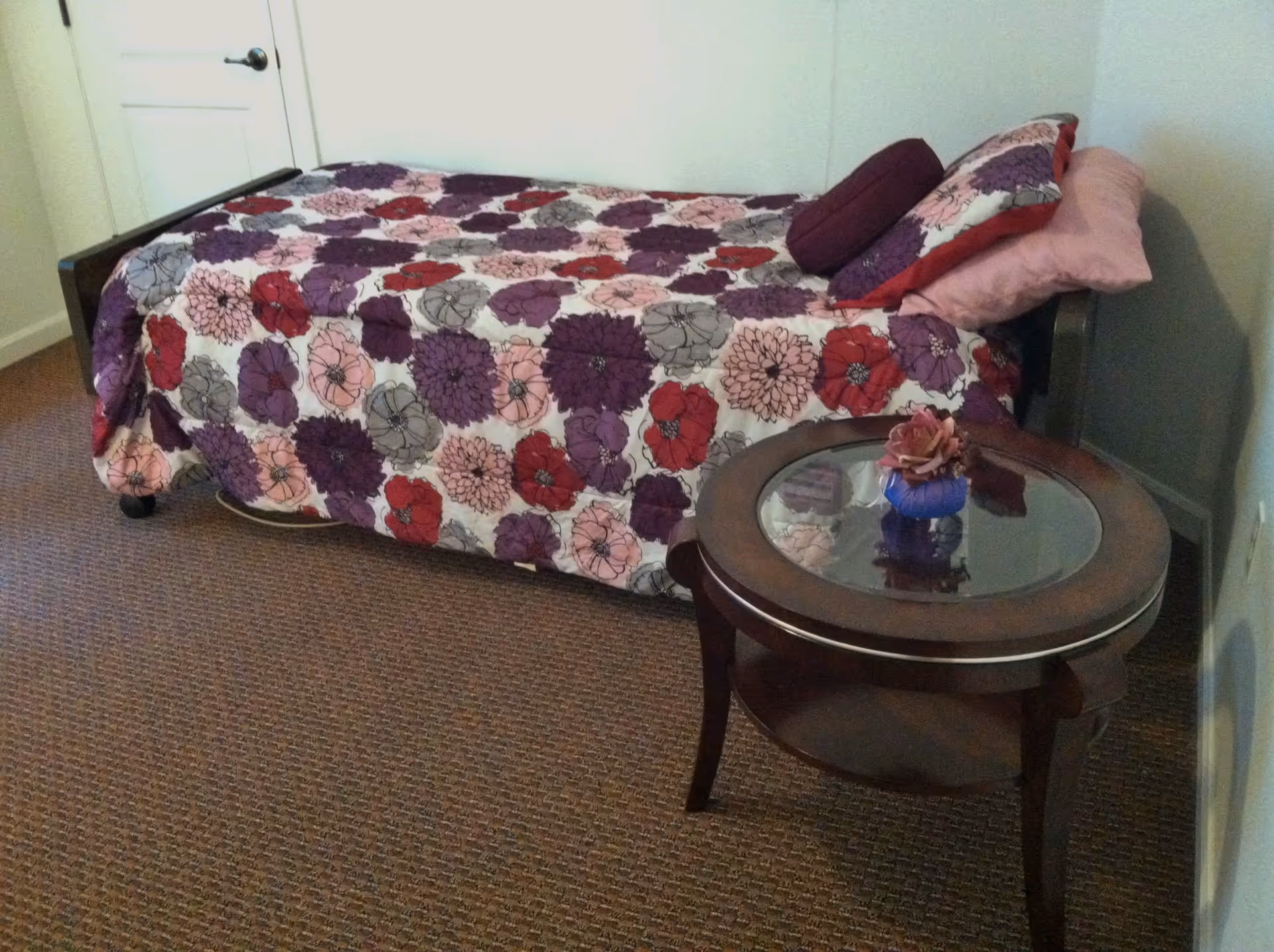 A small bedroom with a single bed covered in a floral patterned bedspread in shades of purple, red, pink, and gray. The bed has matching pillows and is positioned against a white wall. Next to the bed is a round wooden side table with a glass top, holding a small decorative flower arrangement. The floor is carpeted in a brown pattern, and a closed white door is visible in the background.